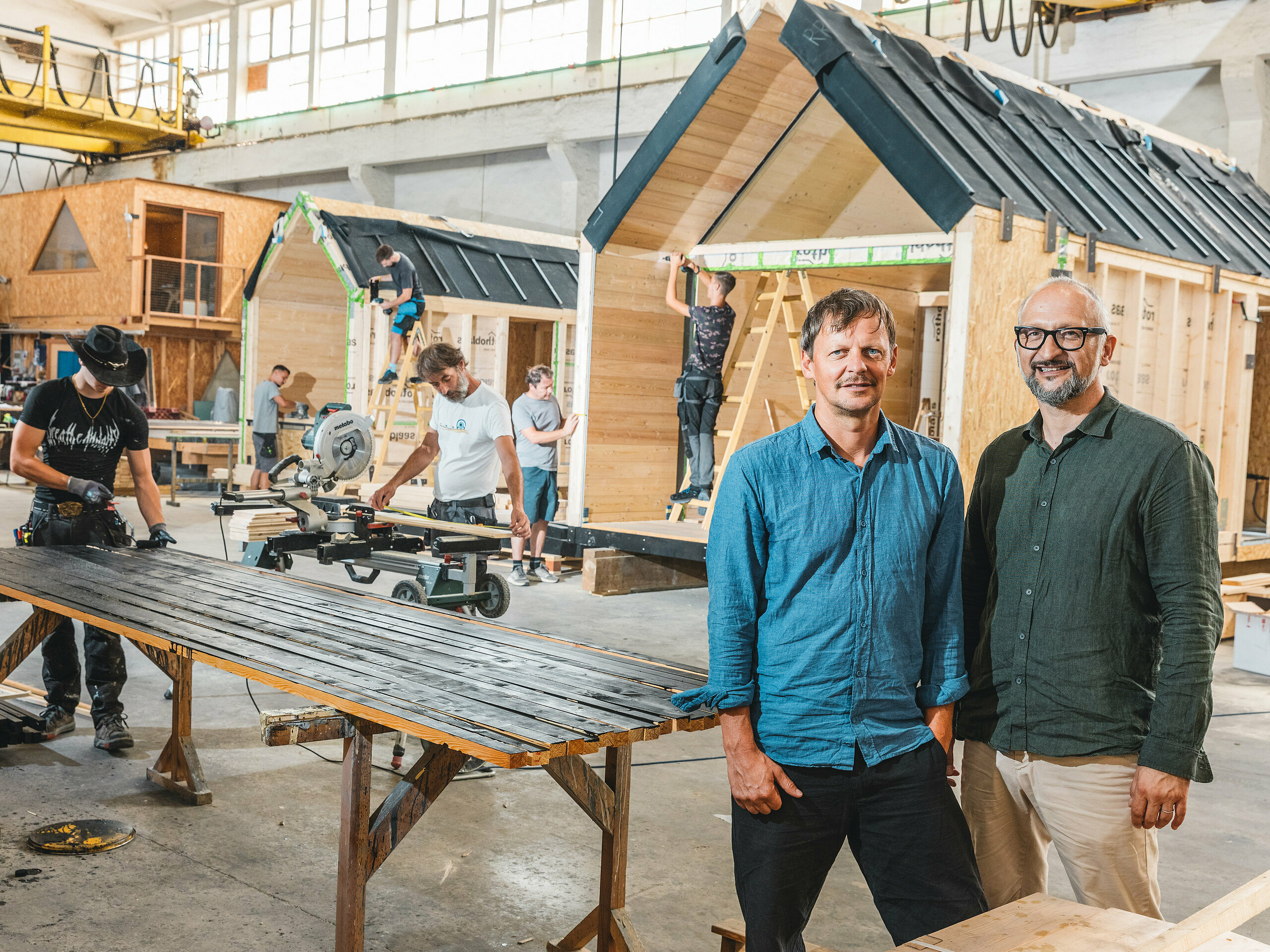 Minimalistické chatky od Lushny se staví ve výrobní hale. Mezinárodně úspěšná společnost se specializuje na miniaturní architekturu pro přírodní resorty. V popředí jsou dva zakladatelé společnosti, architekt Peter Ličen a marketingový specialista Jaka Ažman. V pozadí se montují domy ve tvaru písmene A. Výrazný kontrast dřeva a tmavého hliníku činí z architektury vizuální poutač, o čemž svědčí i Tajná zahrada koi kaprů u jezera Natterer.