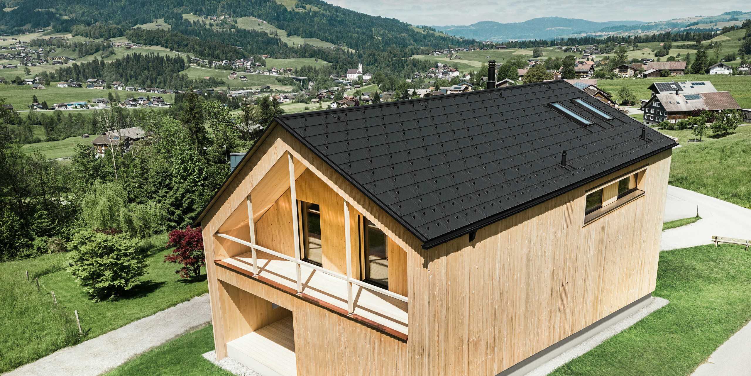 Dieses Foto zeigt ein ökologisches Vorzeigehaus in Egg, das sich mit seiner PREFA Solardachplatte und den schwarzen Dachplatten R.16 gegen die malerische Kulisse eines österreichischen Dorfes abhebt. Die Solarelemente sind nahtlos in die Dachstruktur eingebettet und liefern Energie aus einer erneuerbaren Energiequelle, während das natürliche Holz der Fassade und die sattgrüne Umgebung eine Verbindung von Nachhaltigkeit und Lokalität darstellen. Der Blick auf die umliegende dörfliche Struktur mit traditionellen Bauten im Hintergrund verleiht dem Bild eine zusätzliche Dimension und betont die gelungene Integration moderner, umweltfreundlicher Bauweisen in gewachsene Landschaften.