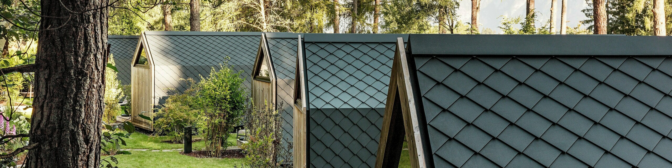 Několik minimalistických domků Tajné zahrady Koi je harmonicky uspořádáno podél zelené zahradní osy u jezera Natterer. Střechy a fasády jsou kompletně obloženy PREFA  fasádními šablonami 20 x 20 v antracitové barvě P.10, které vytvářejí jednotný povrch připomínající rybí šupinu. Obklopené stromy a keři, odolné a povětrnostním vlivům odolné hliníkové produkty PREFA spojují precizní architekturu s klidem, přírodou a vysoce kvalitním glampingovým designem.