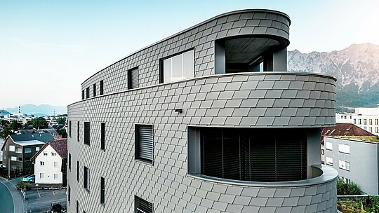 Detailní šikmý pohled na Bernerhaus v Buchsu s jemně zaobleným rohem a souvislými balkonovými pásy. Fasáda, obložená fasádními šindeli PREFA v barvě P.10 světle šedé, překrývá trojúhelníkovou konstrukci jako precizně vyrobený hliníkový plášť a vytváří zářivý, šupinovitý vzor. Homogenní povrch zdůrazňuje plynulé poloměry a hru světla a stínu. Díky lehkému, povětrnostním vlivům odolnému a trvanlivému hliníku PREFA je výsledkem nenáročný obálkový plášť budovy s výrazným vizuálním dopadem na alpské pozadí.