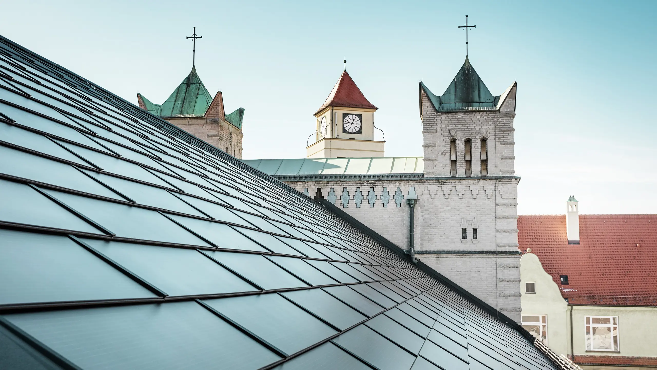 Das prägnante Dach der St. Karl Borromäus Kirche in Nürnberg wurde im Zuge einer umfassenden Sanierung mit schwarzen PREFA-Solardachplatten bedeckt. Der Detailblick auf das Satteldach und die markanten Kirchtürme im Hintergrund vermittelt die gelungene Einbindung moderner Solartechnologie in die historische Architektur der Kirche. Die Solardachplatten sorgen für nachhaltige Energie und fügen sich nahtlos in das traditionelle Erscheinungsbild ein. Dies ist ein weiteres Beispiel für die gelungene Verbindung von Denkmalschutz und innovativer Energiegewinnung. Im Hintergrund ist ein strahlend blauer Himmel zu erkennen.