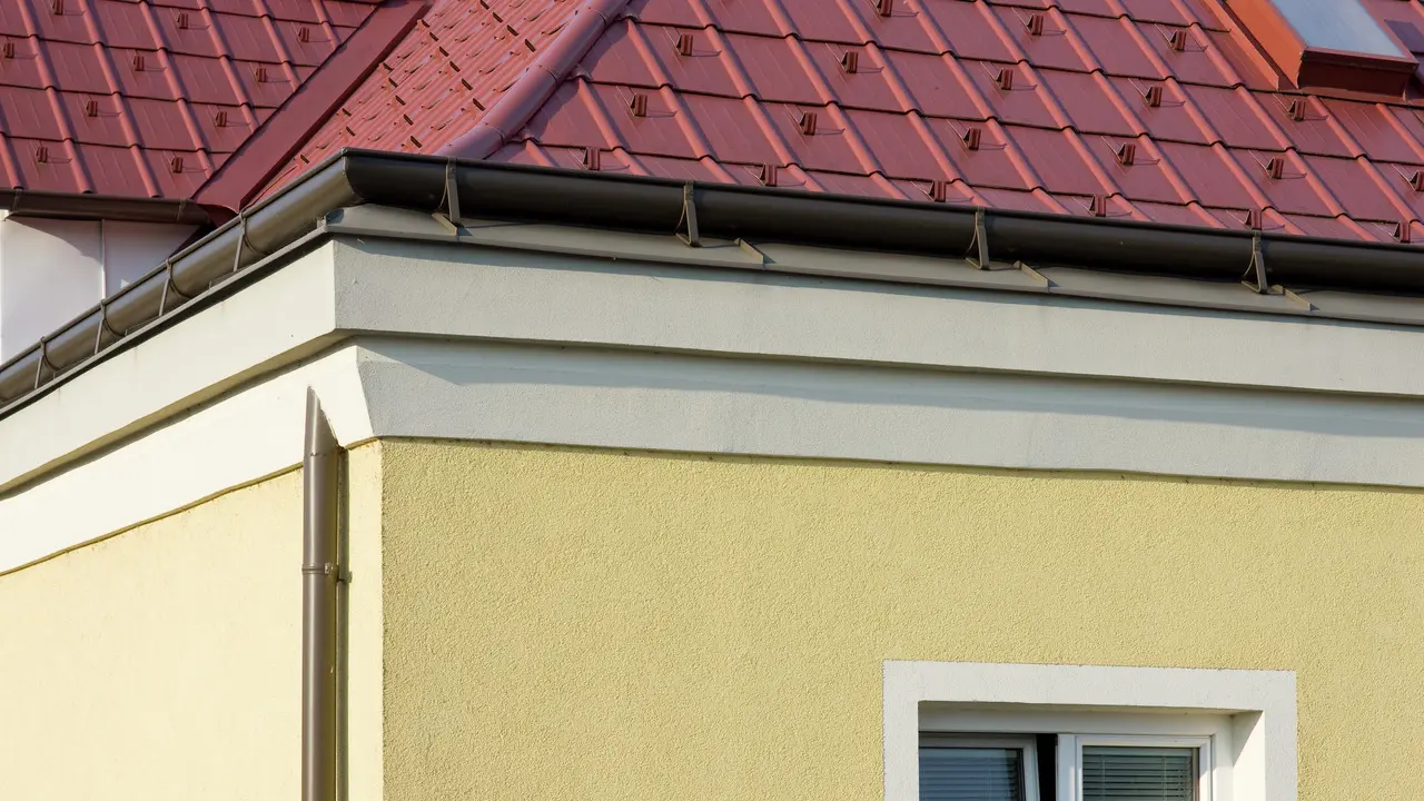 Nároží domu se žlutou omítkovou fasádou a hnědým nástřešním žlabem PREFA s červenou střechou z PREFA falcovaných tašek