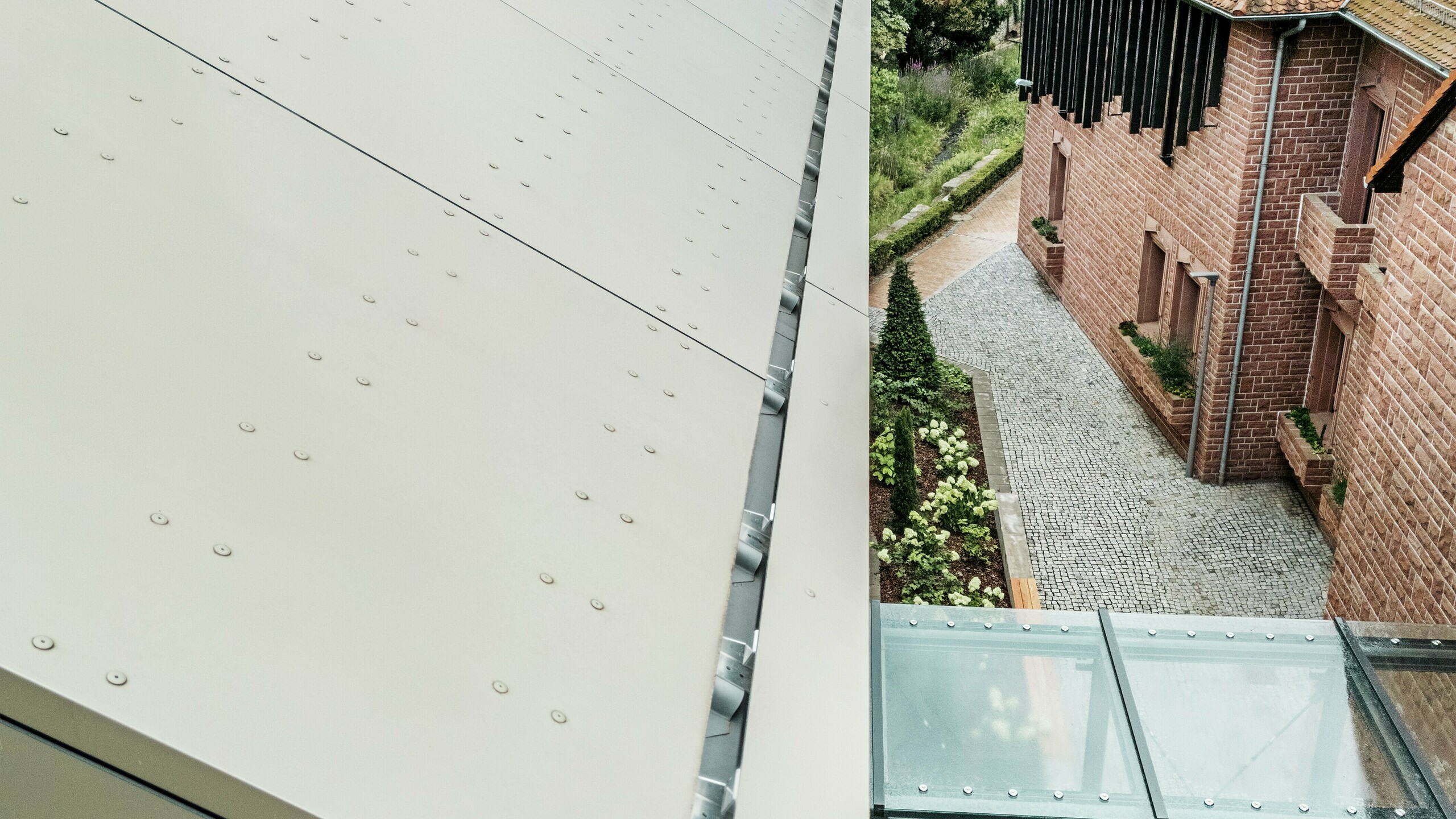 Detailní záběr na střechu radnice v Alzenau, pokrytou hliníkovými kompozitními panely PREFABOND v jemném bronzovém odstínu. Panely jsou precizně namontovány a mají viditelné spojovací prvky, které dodávají fasádě moderní, industriální vzhled. V popředí je vidět skleněná lávka spojující radnici se sousední budovou.