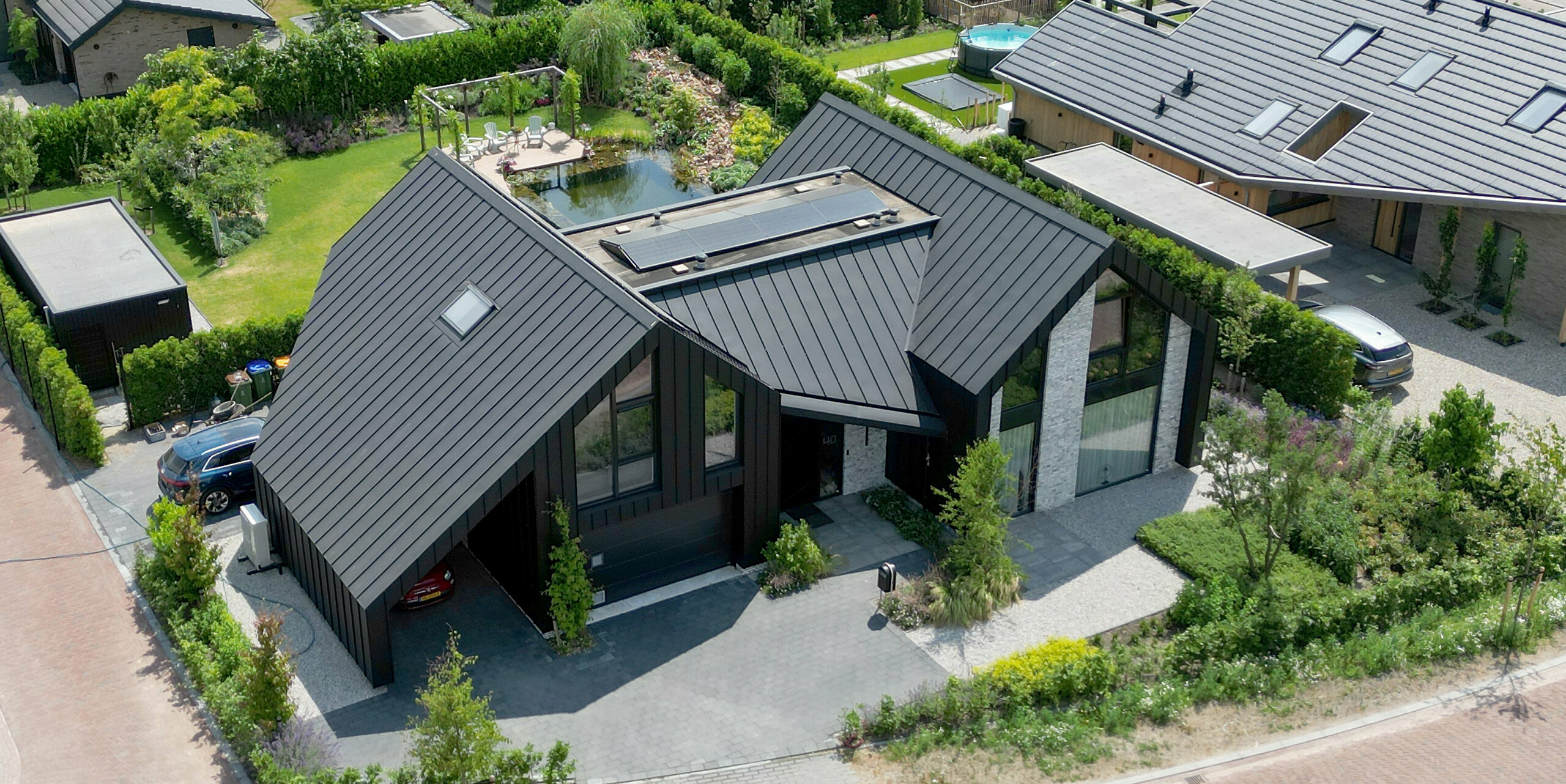 Pohled z ptačí perspektivy na impozantní rodinný dům v nizozemském Veenendaalu, který se vyznačuje černou střechou PREFALZ a harmonickým začleněním do okolní zahradní krajiny. Dům má moderní konstrukci sedlové střechy a je ukázkovým příkladem moderní architektury díky svým jasným liniím a velkým proskleným plochám, orámovaným živou zelení. Dobře promyšlený krajinářský design, včetně soukromé příjezdové cesty a zahrady, umocňuje promyšlenou estetiku nemovitosti.