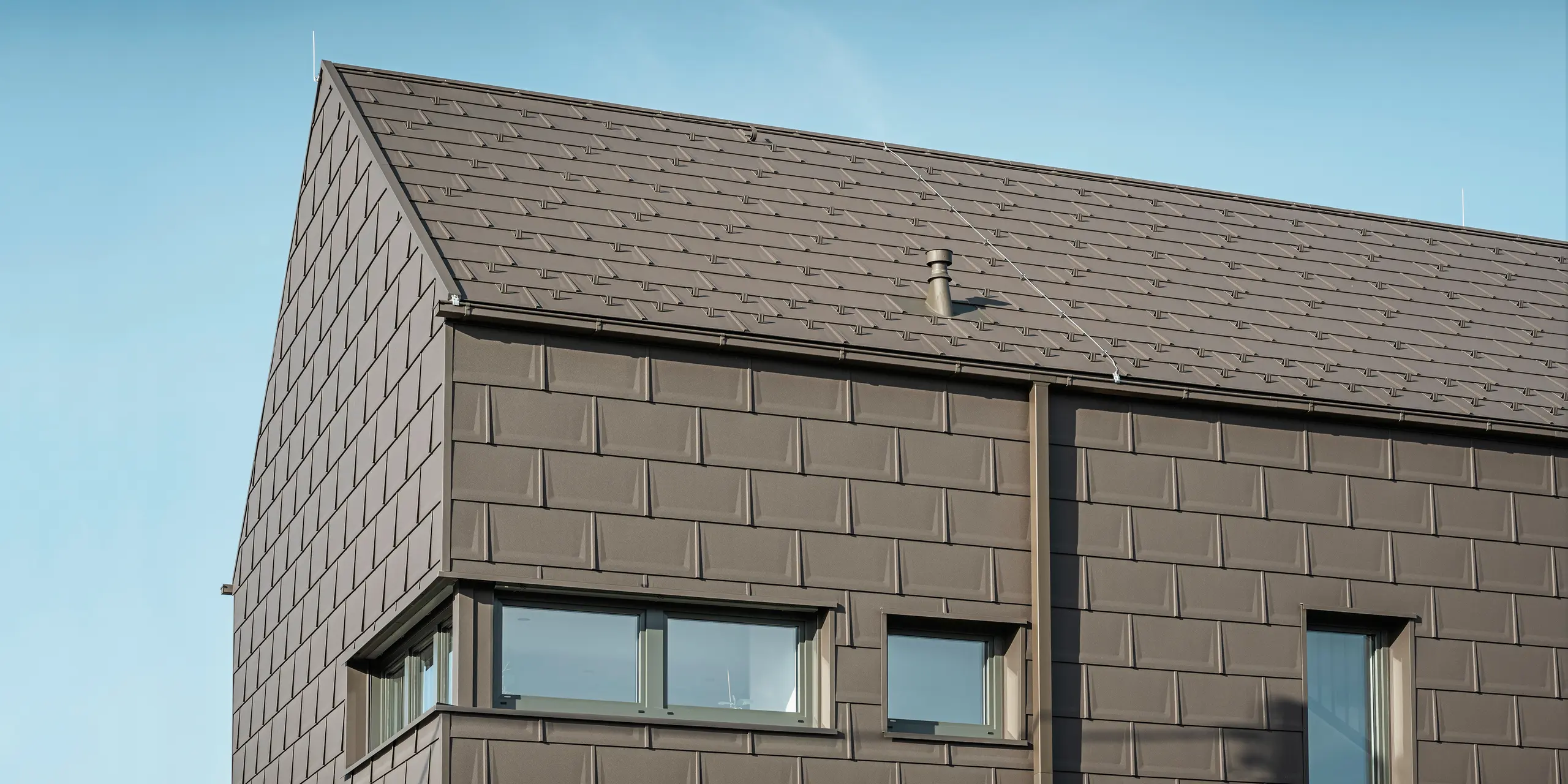Detailní pohled na rodinný dům v rakouském Neukirchenu se střešním a fasádním pláštěm ze střešních panelů PREFA R.16 v P.10 hnědé. Precizní přesah panelů zajišťuje jednotný a atraktivní vzhled, který kombinuje moderní estetiku s funkčností. Integrované střešní odvodnění PREFA v podobě krabicového žlabu a čtyřhranné trubky v hnědé P.10 ukazuje závazek PREFA k vysoce kvalitním a vzhledově atraktivním stavebním dílům.