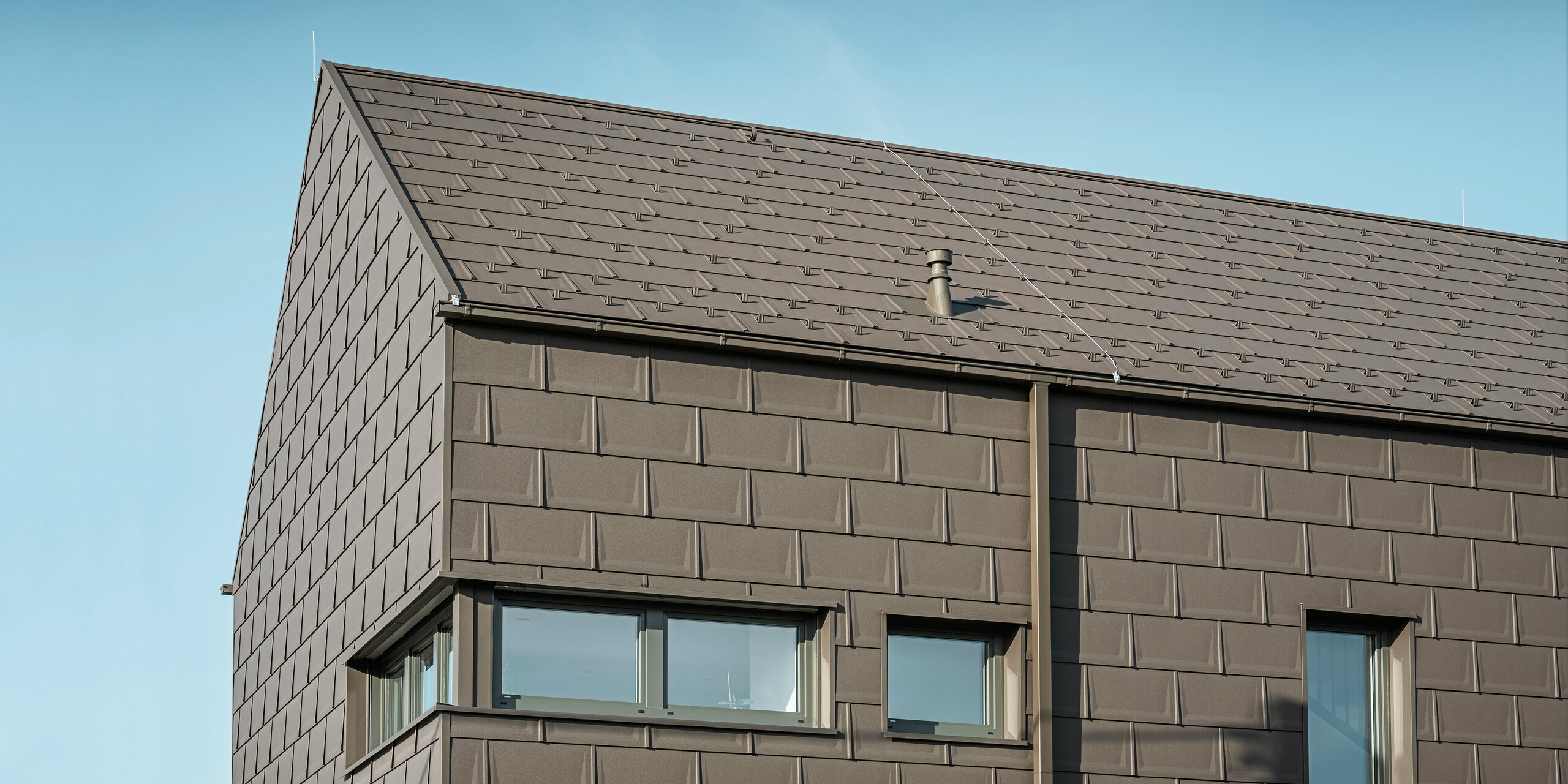 Detailní pohled na rodinný dům v rakouském Neukirchenu se střešním a fasádním pláštěm ze střešních panelů PREFA R.16 v P.10 hnědé. Precizní přesah panelů zajišťuje jednotný a atraktivní vzhled, který kombinuje moderní estetiku s funkčností. Integrované střešní odvodnění PREFA v podobě krabicového žlabu a čtyřhranné trubky v hnědé P.10 ukazuje závazek PREFA k vysoce kvalitním a vzhledově atraktivním stavebním dílům.