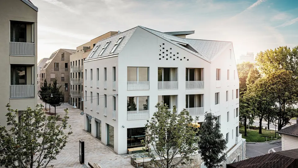 Moderní rezidenční čtvrť Vydrica v Bratislavě se vyznačuje čistým architektonickým stylem a klidnou uliční krajinou. Strmé sedlové střechy s výraznými světlíky jsou pokryty hliníkovými střešními šindeli PREFA v barvách P.10 Prefa bílá, bronzová a P.10 písková, které vytvářejí elegantní plochu na slunci. Precizně provedené linie a homogenní povrchy podtrhují vysoce kvalitní design. Hliník PREFA je známý svou trvanlivostí, nízkou hmotností a vysokou odolností vůči povětrnostním vlivům – ideální pro složité tvary střech a udržitelný, nenáročný plášť budovy.