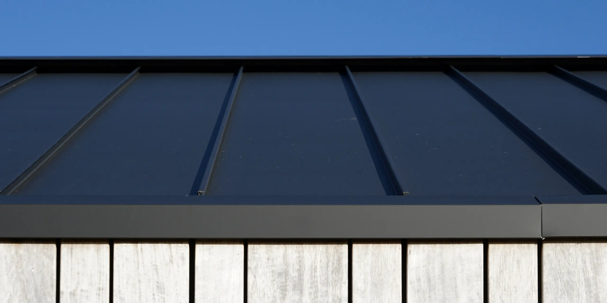 Detailní pohled na moderní rodinný dům v nizozemském Uithoornu s drážkovanou krytinou PREFALZ v černo-šedé barvě. Na obrázku vidíme precizní zpracování vysoce kvalitního plechu PREFA v kombinaci s přírodními dřevěnými prvky na fasádě. 