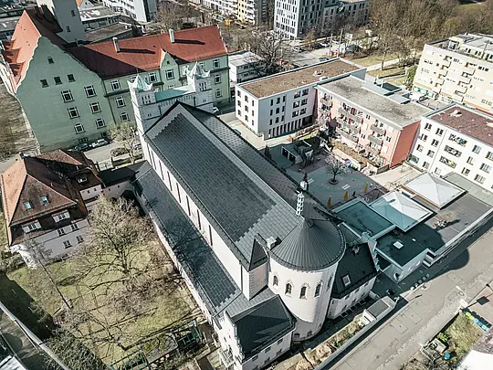 Vogelperspektive auf die Rückseite der St. Karl Borromäus Kirche in Nürnberg. Die schwarzen PREFA Solardachplatten decken das markante, runde Satteldach über dem Hauptteil der Kirche als auch das Dach des Nebengebäudes. Die Aluminiumdachplatten mit integrierten PV-Modulen fügen sich perfekt in das historische Erscheinungsbild der Kirche ein. Der Blick auf das moderne Umfeld mit angrenzenden Wohngebäuden und dem grünen Hofbereich verdeutlicht die gelungene Verbindung von Denkmalschutz, moderner Architektur und nachhaltiger Energiegewinnung. Eine ideale Lösung für den Denkmalschutz im urbanen Raum.