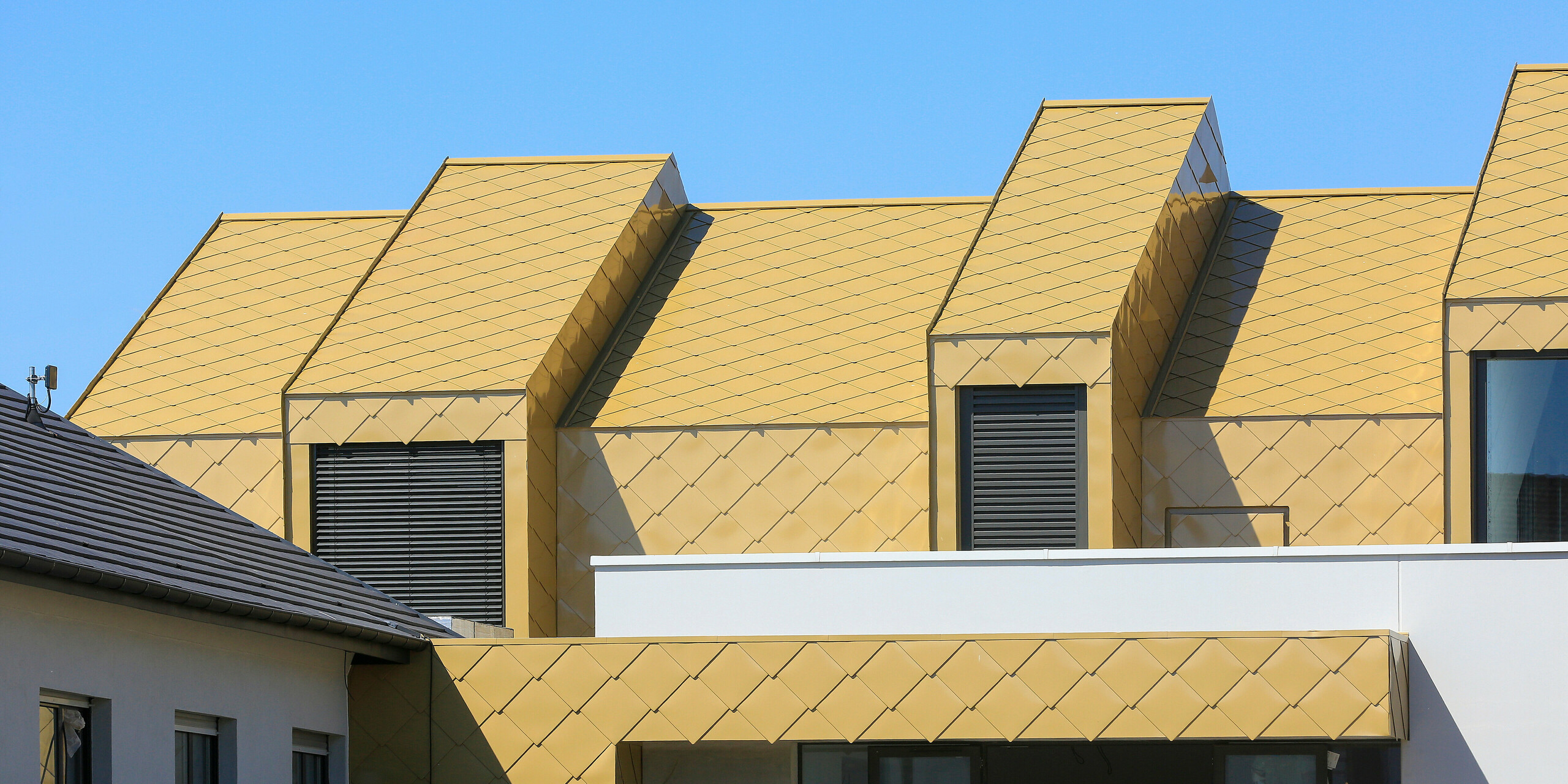Detailní záběr na balkon a střešní okna objektu ve Fessenheim. Střecha a části fasády byly pokryty falcovanými a fasádními šablonami PREFA 44 × 44 v barvě Sparkling Gold.