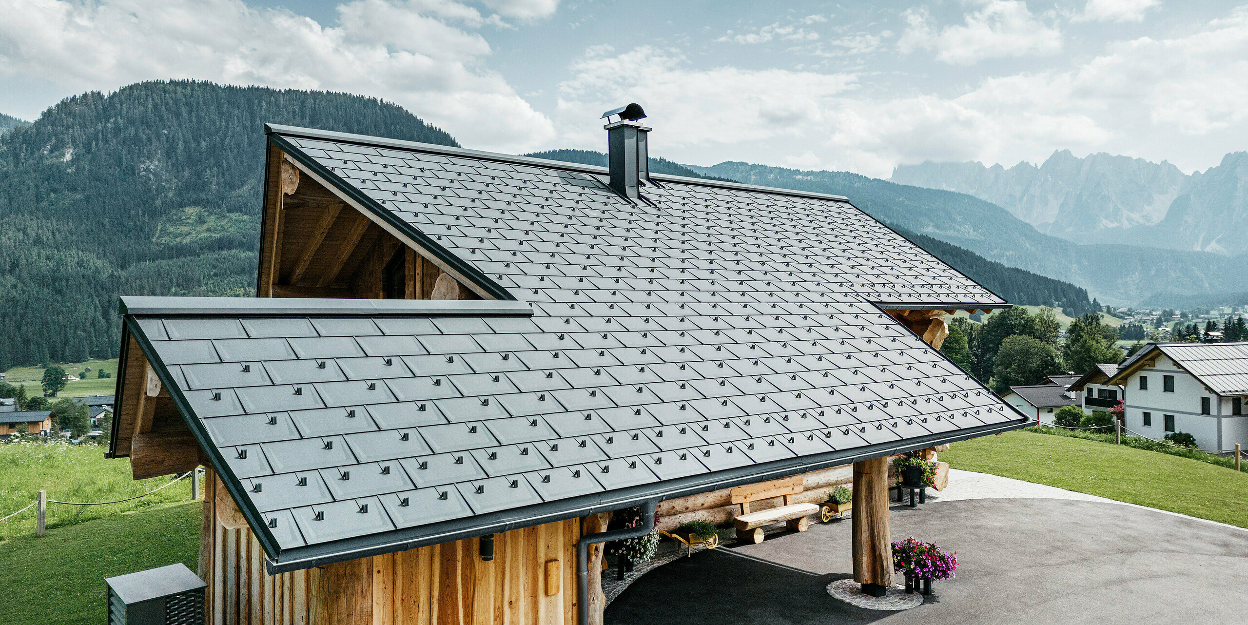 moderní a zároveň tradiční architektura spojená ve srubovém domě v rakouském Gosau. Dřevěný dům má střechu ze střešních panelů PREFA R.16 v barvě P.10 antracitové. Letecký pohled ukazuje preciznost střešní konstrukce, dokonalé začlenění do designu srubu a idylické začlenění do alpské krajiny s úchvatným horským panoramatem v pozadí. Střešní panely dodávají domu současný nádech a jsou v souladu s ekologicky šetrnou a udržitelnou výstavbou.