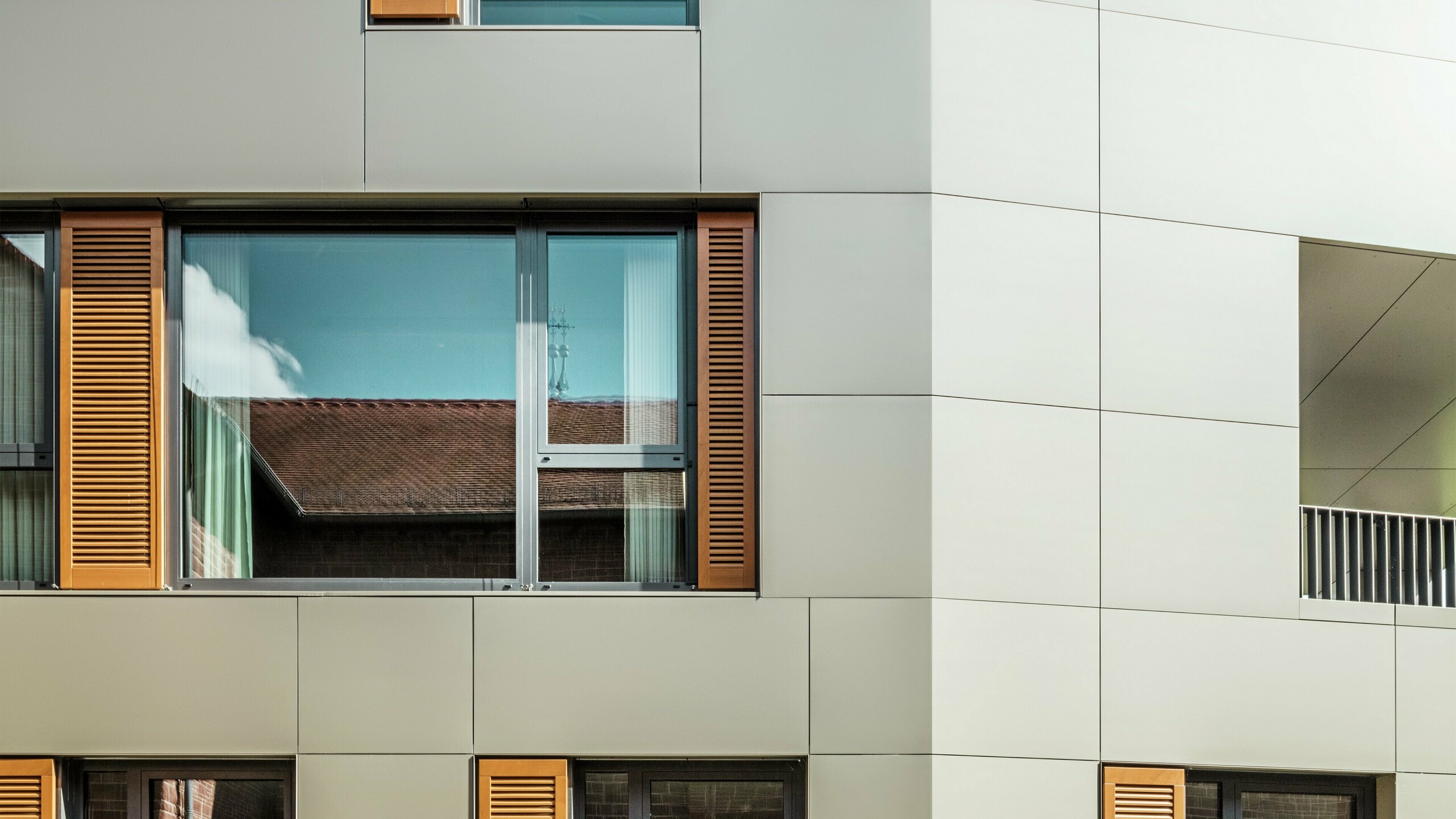 Detailní snímek fasády radnice v německém Alzenau s bronzovými hliníkovými kompozitními panely od společnosti PREFA. Velká okna jsou orámována elegantními dřevěnými žaluziemi v teplé hnědé barvě, které fasádě dodávají atraktivní texturu. V popředí doplňuje udržovaný květinový záhon městský design budovy zelenými akcenty. Jasná obloha a čisté linie architektury podtrhují moderní charakter budovy.