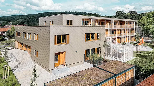 Z výšky je vidět novostavba denní péče v Alzenau ve tvaru písmene L s přilehlým obytným křídlem – progresivní hybridní budova postavená s využitím udržitelných metod. Bronzově zbarvená, zavěšená provětrávaná hliníková fasáda PREFA s fasádními šablonami o rozměrech 44 x 44 cm definuje moderní, strukturovaný exteriér denní péče. Velkorysá dřevěná okna a vertikální žaluzie vytvářejí příjemnou a teplou atmosféru. Zadní část s venkovním schodištěm, hracími plochami a zelenými plochami ilustruje funkční oddělení a současné propojení různých využití. Zelená střecha a kombinace materiálů – hliníku, dřeva a betonu – odrážejí vysoké ekologické standardy projektu a jsou silným poselstvím pro udržitelnou vzdělávací a bytovou architekturu.