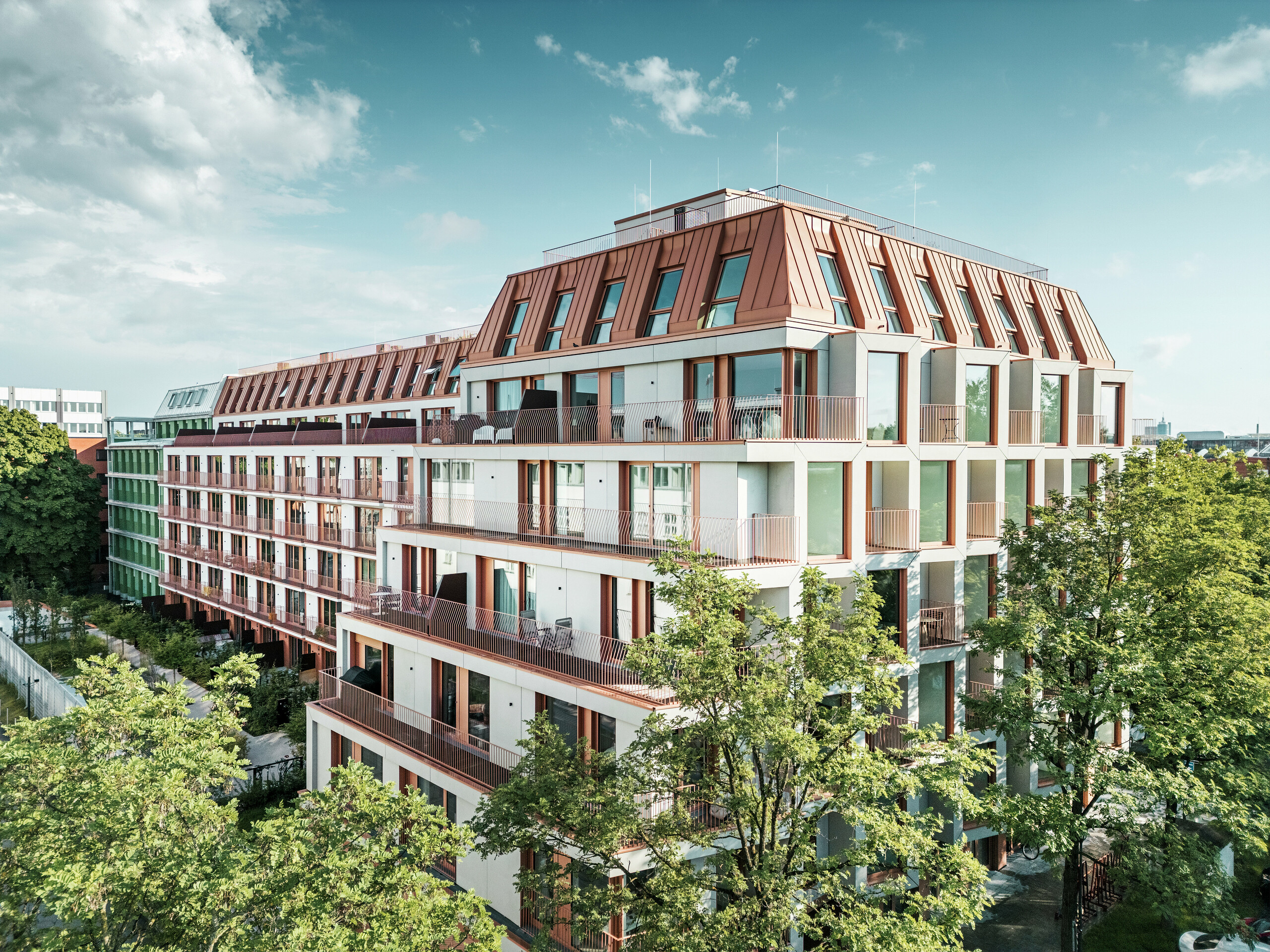 Celkový pohled na městský rezidenční projekt Van B v Mnichově s čistou architekturou, zelenými venkovními plochami a výraznou hliníkovou střechou PREFALZ z IGP Orange HDP od PREFA. Odolný a nenáročný střešní materiál tvoří jednotící designový prvek budovy a zajišťuje dlouhodobé a estetické řešení v městském prostoru. Velkorysklá okna, balkony a promyšlené dispozice nabízejí moderní komfort bydlení s vysokou kvalitou designu.