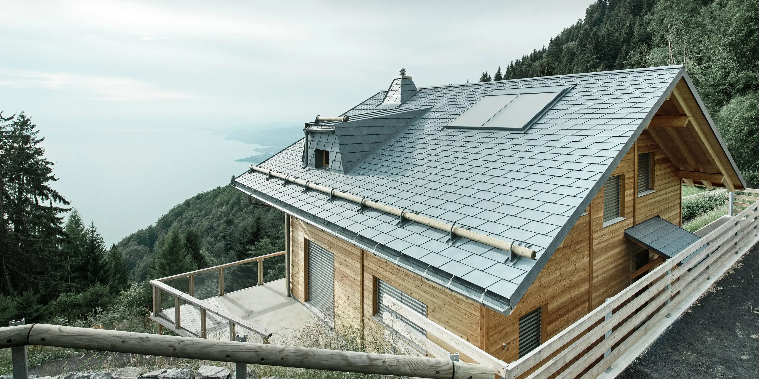 Dachansicht einer Holz-Lodge am Genfersee mit PREFA Dachschindeln in P.10 Steingrau. Die präzise verarbeitete Dachgaube und der Kamin in Trapezform setzen markante Akzente, während das robuste Dach Wind und Wetter trotzt. Das elegante Design aus Aluminium in Kombination mit Holz sorgt für eine perfekte Harmonie zwischen Funktion und Ästhetik. Im Hintergrund ist der in mystischen Nebel gehüllte Genfersee zu erkennen.