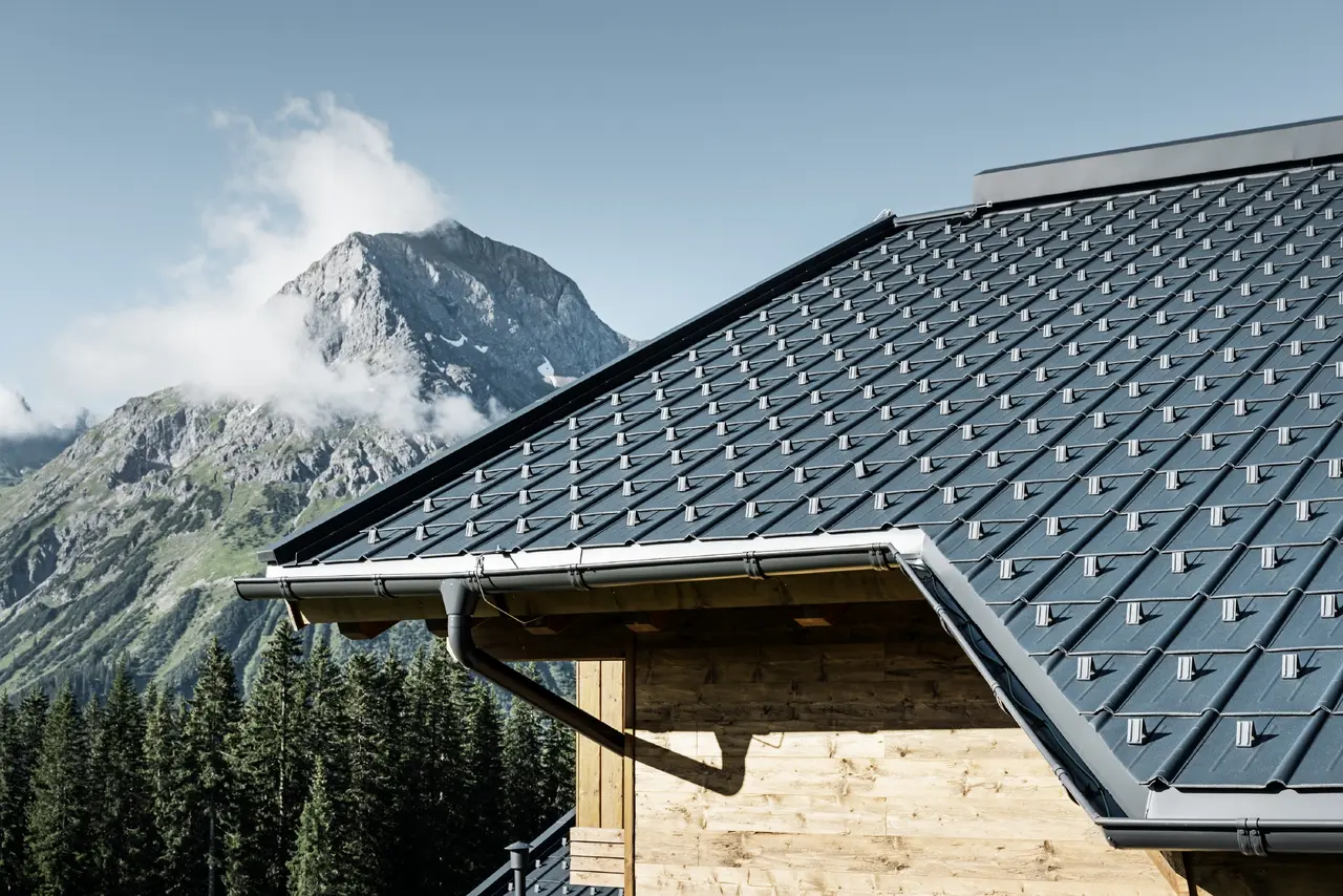 PREFA Dachplatte im alpinen Einsatz - Dachplatte in Anthrazit verlegt auf Hütte in Lech am Arlberg mit Berge im Hintergrund; PREFA Dachrinne in Anthrazit; Holzfassade