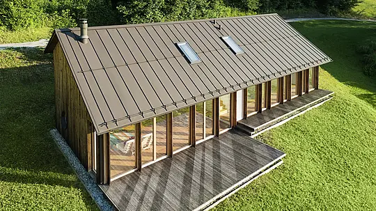 Moderní dřevěný dům s velkorysou prosklenou fasádou a odolnou hliníkovou střechou PREFALZ v barvě P.10 tmavě – ideální pro svažité lokality, jako je právě Vinje. Vysoce kvalitní střešní krytina zajišťuje maximální odolnost vůči povětrnostním vlivům, dlouhou životnost a elegantní vzhled. Přesná montáž stojatých drážek a integrovaných střešních oken vytvářejí prosvětlený a udržitelný koncept bydlení. Pohled z ptačí perspektivy demonstruje precizní zpracování této drážkové krytiny.