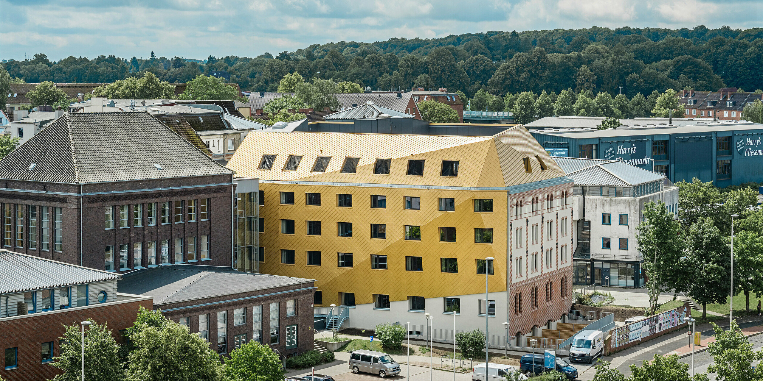 Blick aus der Ferne auf "The Station" in Kiel mit schönen Stadtpanorama im Hintergrund. Das Studentenwohnheim sticht hervor durch die auffällige Aluminiumfassade sowie das Dach bestehend aus PREFA Dach- und Wandrauten 29 × 29 in Mayagold.