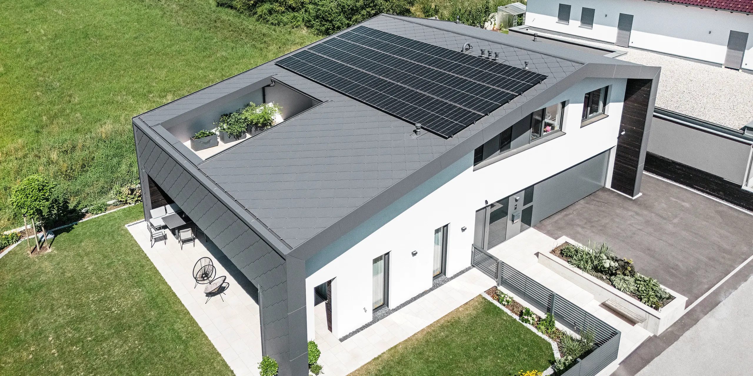 Dronový snímek moderního rodinného domu v Aschachu s tmavě šedými střešními a fasádními šablonami PREFA 44 × 44 v kvalitním povrchu P.10. Rozsáhlá střešní plocha je vybavena fotovoltaickou instalací a atriem. Atraktivní architektura navíc zahrnuje krytou terasu. Střecha a fasáda z hliníku jsou lehké, odolné a poskytují optimální ochranu před povětrnostními vlivy. Přední strana budovy je opatřena bílou omítkovou fasádou.