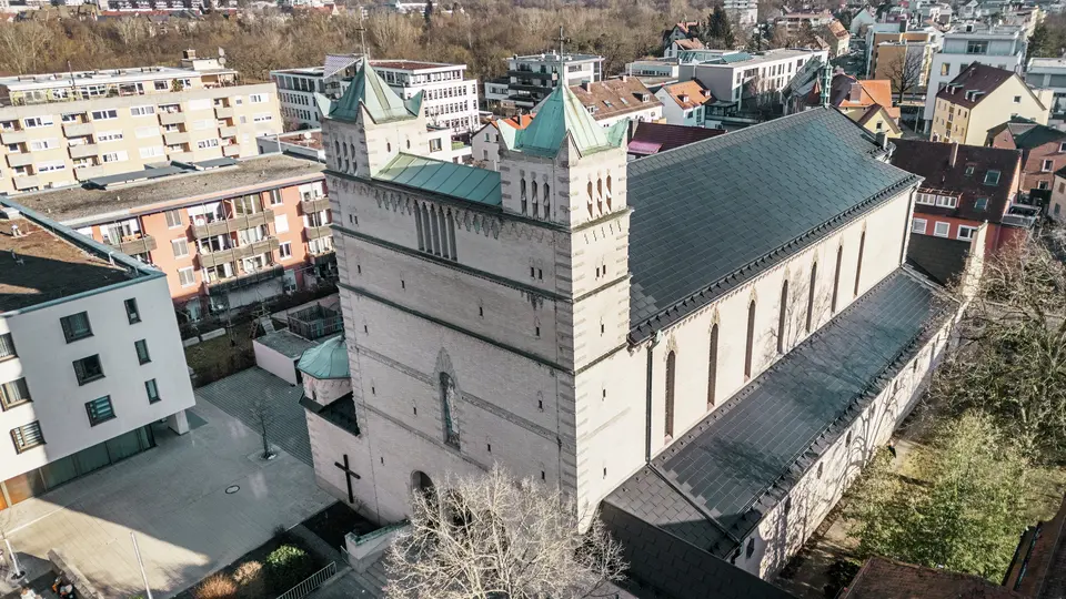 Vorderseite der St. Karl Borromäus Kirche in Nürnberg: Das Bild zeigt das beeindruckende Dach der denkmalgeschützten Kirche, das mit schwarzen PREFA-Solardachplatten gedeckt ist. Die Solarmodule fügen sich harmonisch in die historische Architektur der Kirche ein und stellen gleichzeitig eine moderne Lösung zur nachhaltigen Energiegewinnung dar. Im Hintergrund sind moderne Gebäude und die grünen Hügel Nürnbergs zu sehen, was den gelungenen Übergang zwischen traditioneller Kirchenarchitektur und zeitgemäßer Energieversorgung verdeutlicht. Das Aluminiumdach der Kirche wird durch die stilvolle PREFA Dachplatte R.16 vervollständigt.