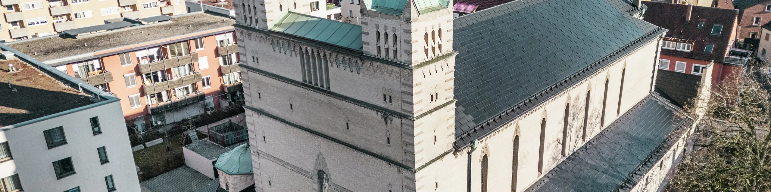 Vorderseite der St. Karl Borromäus Kirche in Nürnberg: Das Bild zeigt das beeindruckende Dach der denkmalgeschützten Kirche, das mit schwarzen PREFA-Solardachplatten gedeckt ist. Die Solarmodule fügen sich harmonisch in die historische Architektur der Kirche ein und stellen gleichzeitig eine moderne Lösung zur nachhaltigen Energiegewinnung dar. Im Hintergrund sind moderne Gebäude und die grünen Hügel Nürnbergs zu sehen, was den gelungenen Übergang zwischen traditioneller Kirchenarchitektur und zeitgemäßer Energieversorgung verdeutlicht. Das Aluminiumdach der Kirche wird durch die stilvolle PREFA Dachplatte R.16 vervollständigt.