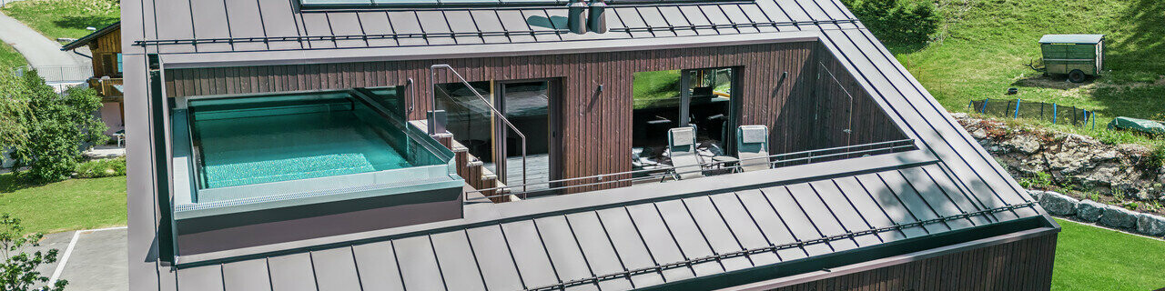 Pohled na moderní bytový dům se střechou od PREFA s Prefalzem v barvě ořechově hnědé. Na obrázku je také terasa s bazénem obklopená dřevěným obkladem a skleněným zábradlím.
