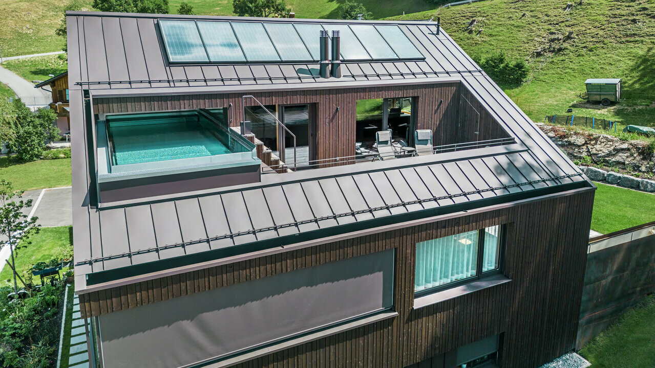 Pohled na moderní bytový dům se střechou od PREFA s Prefalzem v barvě ořechově hnědé. Na obrázku je také terasa s bazénem obklopená dřevěným obkladem a skleněným zábradlím.