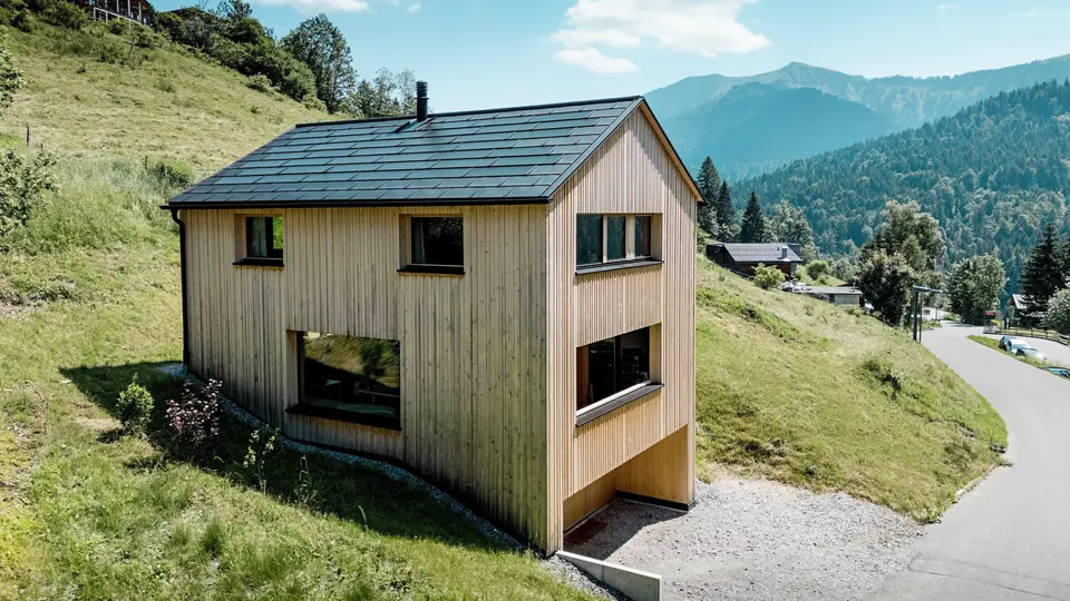 Vysoko nad údolím se na slunném svahu hory Ebnit tyčí dům Haus Immi jako drsný dřevěný monolit. Minimalistická fasáda z bílé jedle a černá solární střecha PREFA vytvářejí výrazný kontrast k rozlehlé alpské krajině. Integrované solární střešní panely a střešní panely R.16 v černé barvě P.10 vyrábějí solární energii, odolávají alpskému počasí a zajišťují dlouhodobý a harmonický vzhled střechy.