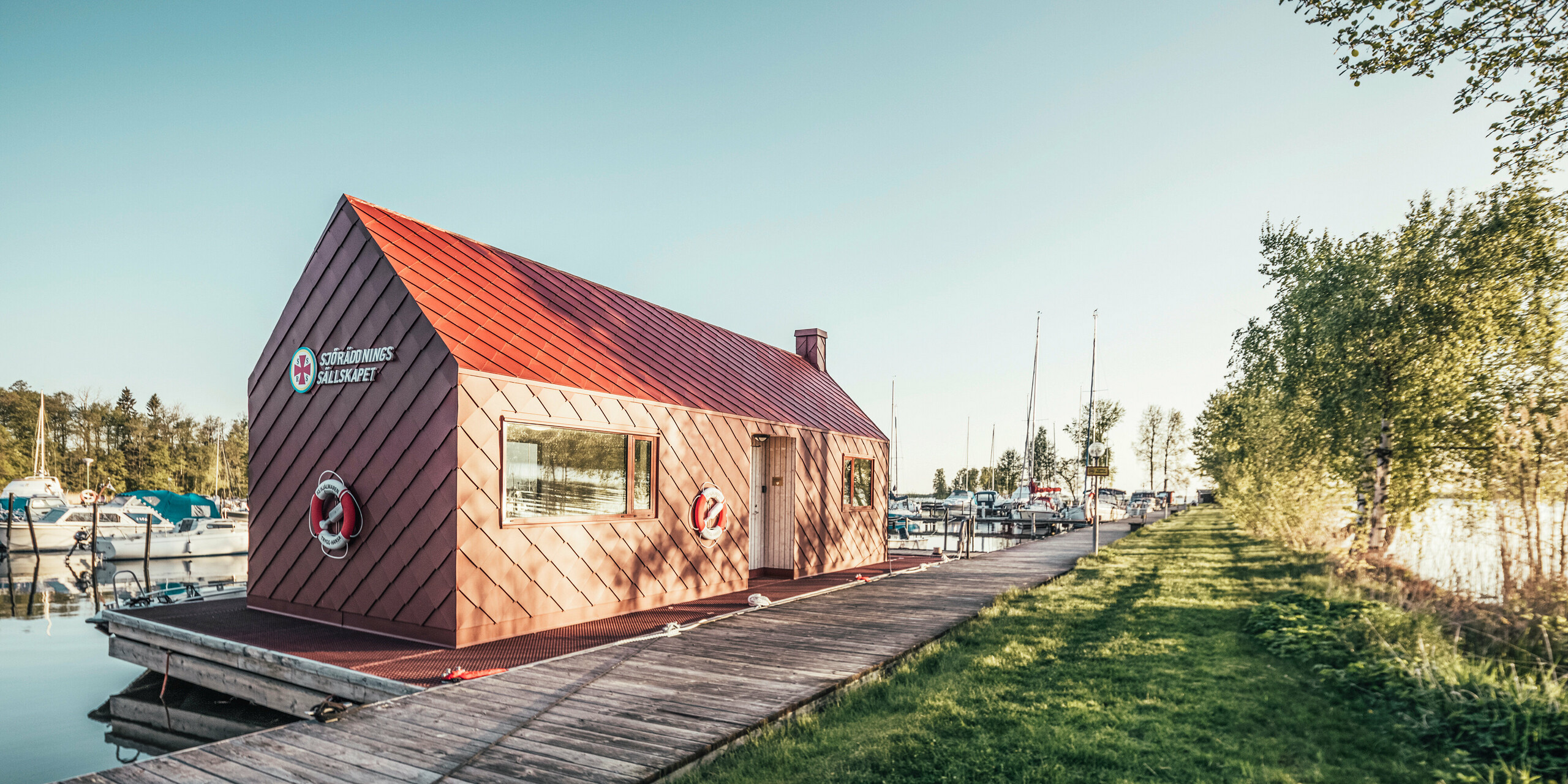 Námořní záchranná stanice v harmonickém prostředí na jezeře Hjälmaren ve Švédsku s tmavě červenou střechou PREFA a fasádními šablonami 29×29. Stanice je na konci dřevěného mola, které vede do přístavu. Zelené stromy rámují obraz, který zachycuje klidnou atmosféru záchranné stanice ve zlatém ranním světle.