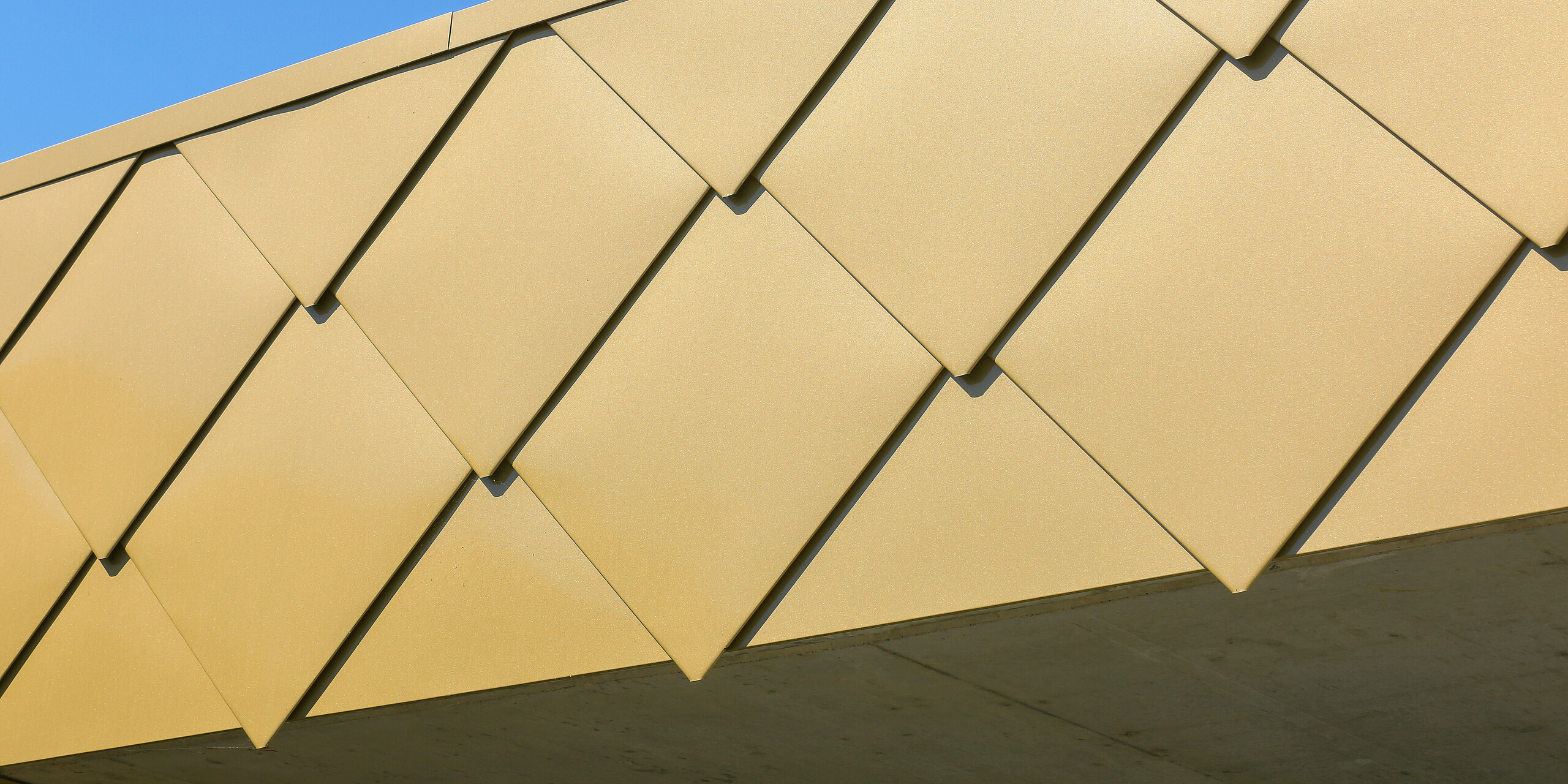 Detailní záběr na lesklé fasádní šablony PREFA 44 × 44 v barvě na přání Sparkling Gold na kulturním centru Fessenheim ve Francii. Díky pozoruhodným hliníkovým šablonám se budova leskne během dne.