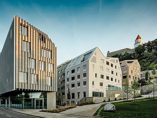 Pozoruhodná souhra moderní architektury a historického kontextu v bratislavské čtvrti Vydrica: V popředí upoutá pozornost sochařská budova s ​​vertikální fasádní strukturou, zatímco za ní strmě šikmé střechy s hliníkovými střešními šindeli PREFA v bílé a pískové barvě vytvářejí precizní akcenty. Jasně strukturované střešní plochy s integrovanými okny tvoří silný vizuální kontrast s kulisou hradu na svahu. Hliník PREFA přesvědčí svou vysokou tvárností, trvanlivostí a extrémní odolností vůči povětrnostním vlivům – ideální pro architektonicky náročné městské projekty.