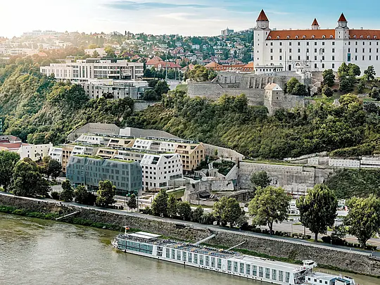 Panoramatický výhled na bratislavskou čtvrť Vydrica s výhledem na hrad a Dunaj: Mnohoforemná architektura zaujme zelenými střechami, strmými šikmými střechami a precizně integrovanými střešními okny jako vizuálně výraznými prvky. Hliníkové střešní šindele PREFA v odstínech prefa bílá, písková a bronzová spojují stavební konstrukce do harmonického celku a plynule zapadají do historického prostředí. Odolná, lehká a extrémně odolná řešení PREFA umožňují složité geometrie střech a zajišťují udržitelný, nenáročný na údržbu plášť budovy s vysokou estetickou kvalitou.