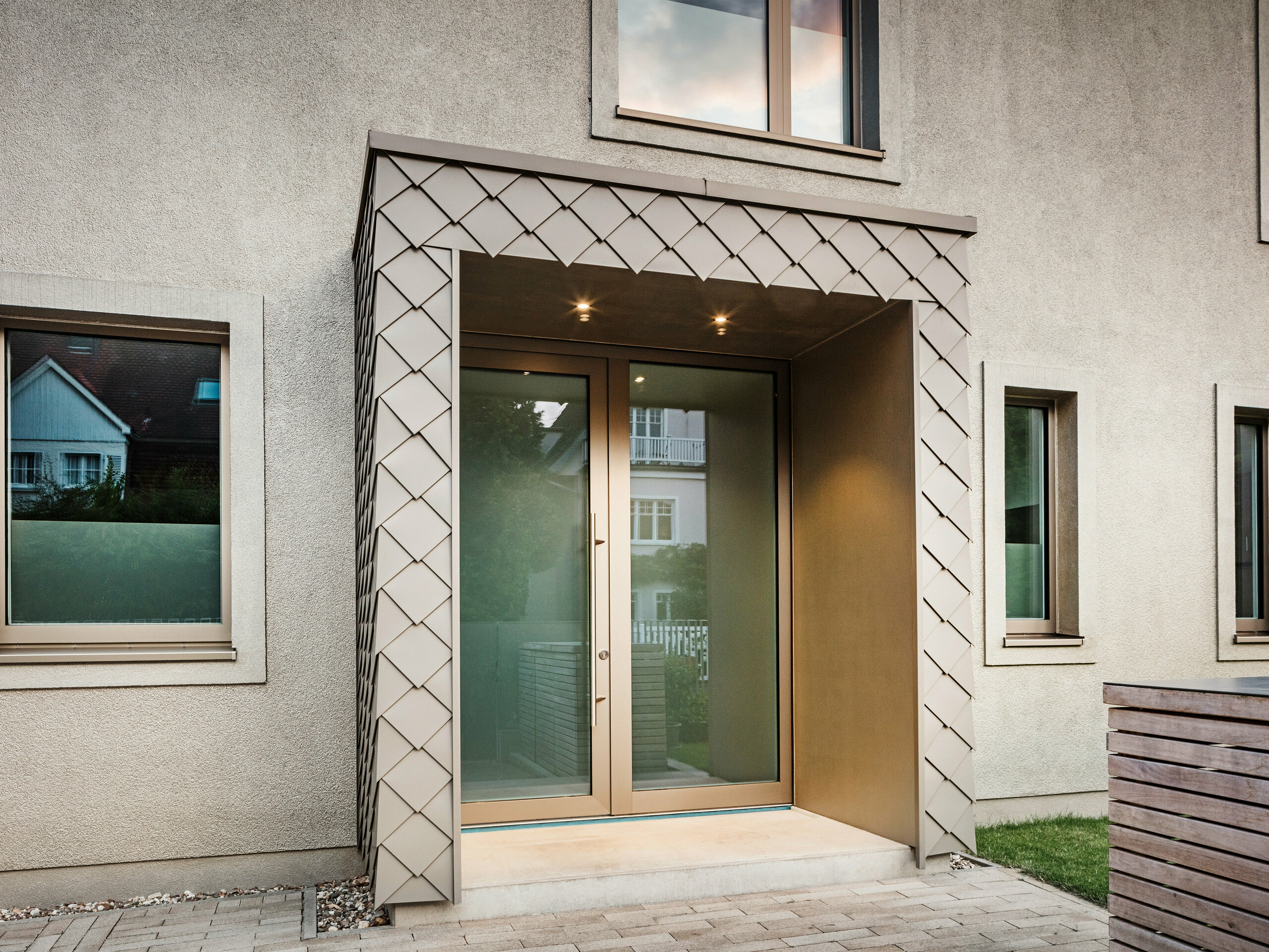 Detailní záběr vstupního prostoru moderního rodinného domu s hliníkovým opláštěním v bronzové barvě od PREFA. Vchod je pokrytý a pokrytý elegantními hliníkovými šablonami, což dává prostoru moderní a stylový vzhled. Nalevo a napravo od vstupních dveří jsou velká okna, která vytvářejí světlou a příjemnou atmosféru.