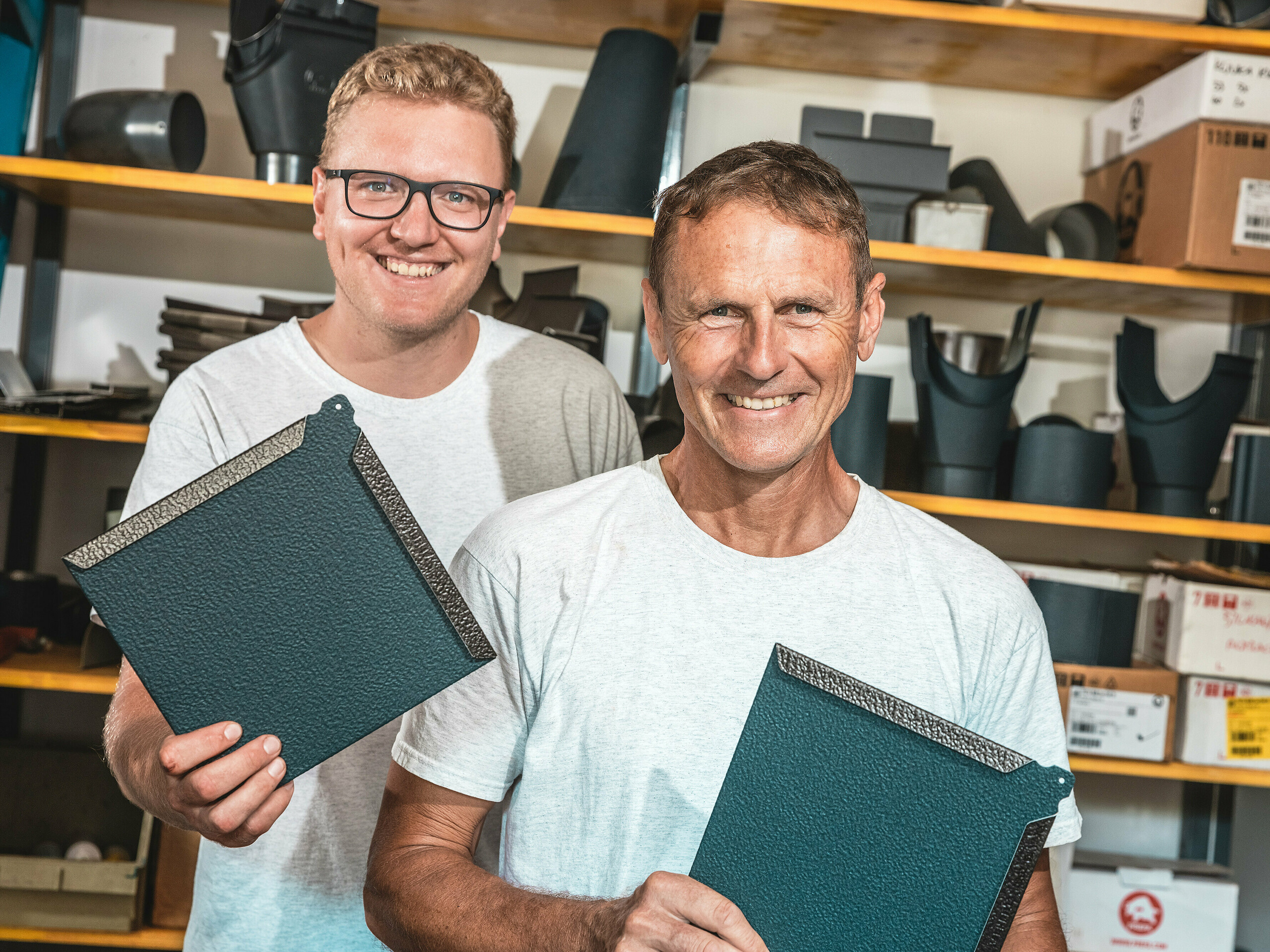 V dílně dva řemeslníci prezentují PREFA fasádní šablony 20 x 20 v antracitové barvě P.10. Muž v popředí je slovinský klempířský mistr Janez Kralj. Za ním stojí mladý člen jeho týmu. V pozadí jsou vidět regály plné komponentů a příslušenství pro střešní a fasádní systémy. Jemně texturovaný povrch hliníkových panelů PREFA svědčí o jejich vysoké kvalitě materiálu, odolnosti a precizním zpracování. Jsou ideální pro náročné architektonické projekty, jako jsou malé domy, kde je nezbytná odolnost, nízká hmotnost a moderní design