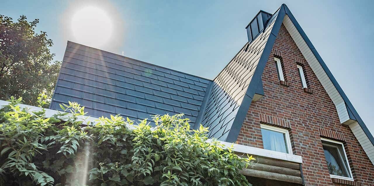 Sedlová střecha pokrytá hladkým hliníkovým střešním panelem PREFA R.16 v antracitové barvě. Fasáda je rustikální cihlová. Nad domem právě vychází slunce.
