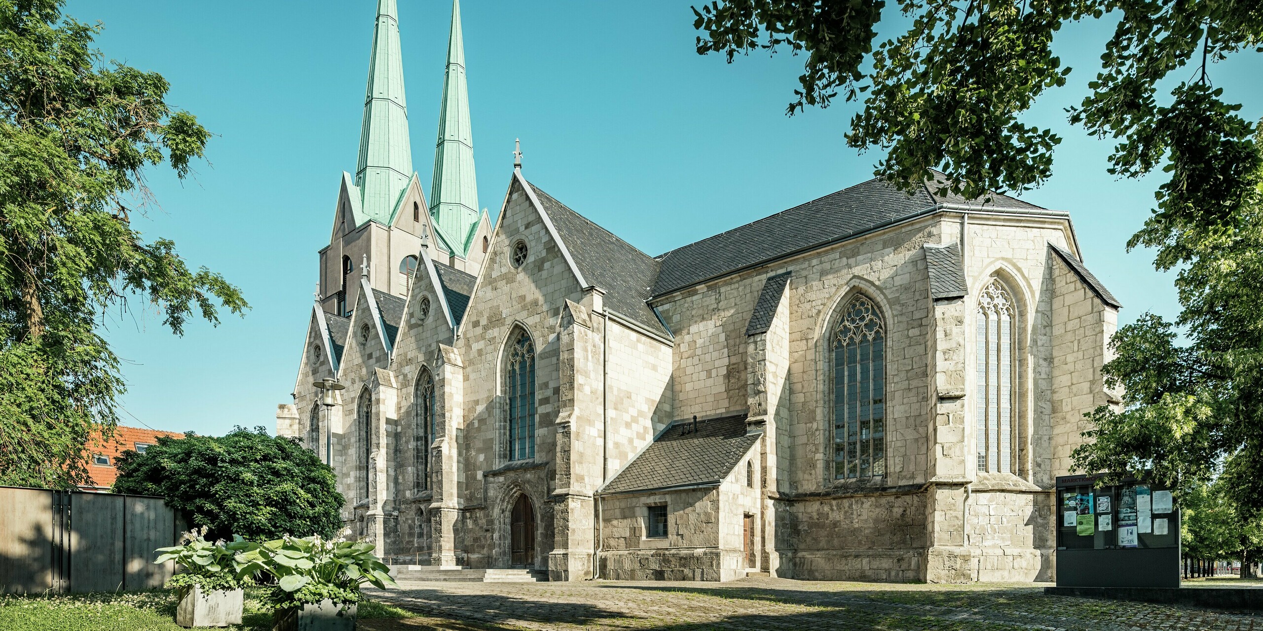 Harmonische Seitenansicht der sanierten Stadtkirche St. Johannis in Ellrich, eingerahmt von Bäumen und strahlend blauem Himmel. Die auffälligen Turmspitzen mit PREFALZ in P.10 Patinagrün bilden einen starken Kontrast zum historischen Natursteinmauerwerk. Das hochwertige Aluminiumdach unterstreicht die moderne Interpretation des Kirchengebäudes und bietet maximale Beständigkeit und Schutz.