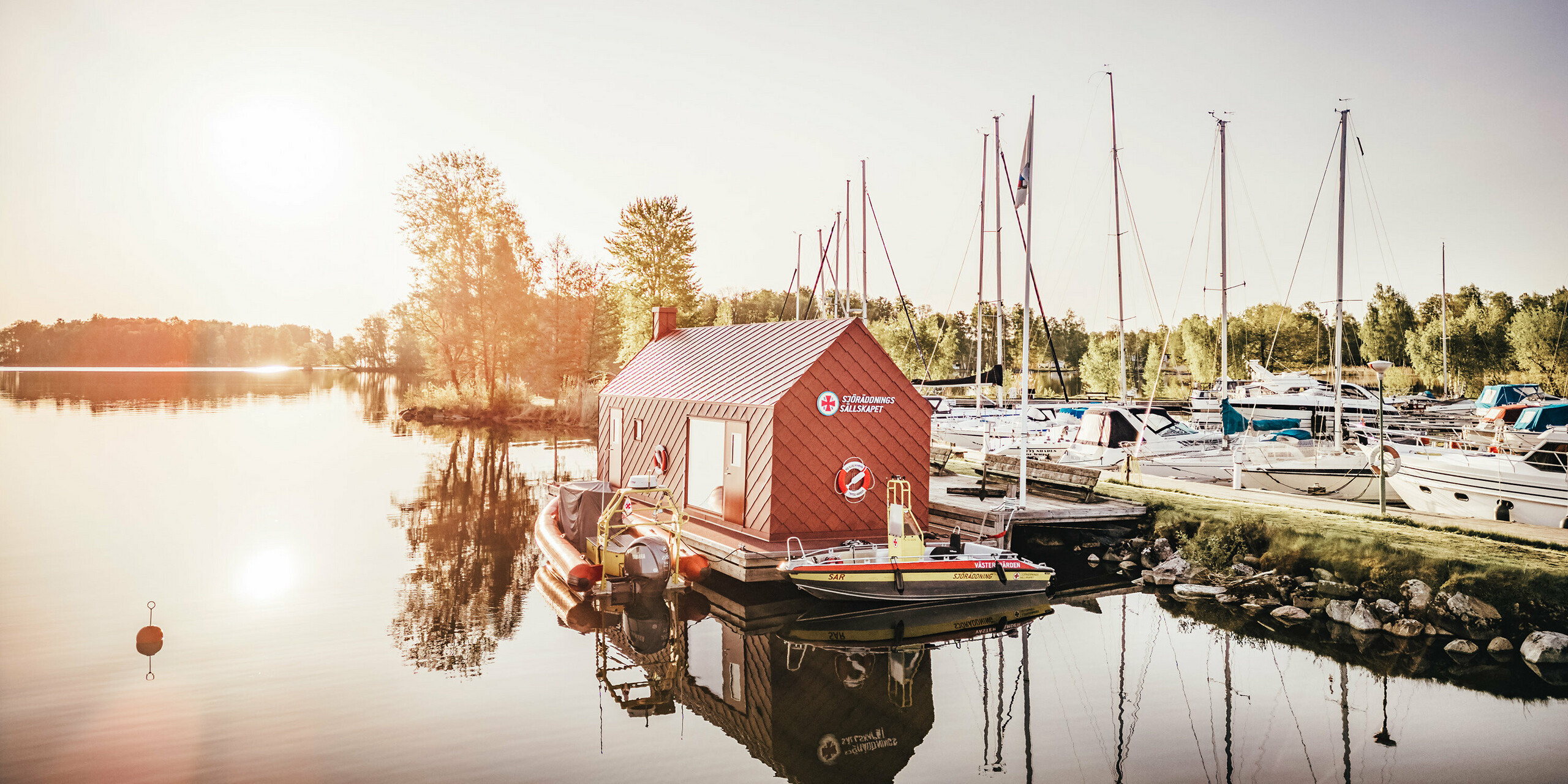 Časně zrána u jezera Hjälmaren ve Švédsku, kde plovoucí dům vodní záchranné stanice s tmavě červeným hliníkovým pláštěm vyrobeným z PREFA hliníkových šablon 29×29 tiše leží na vodě. Vycházející slunce za budovou zalévá scénu měkkým světlem, které se odráží ve stojaté vodě spolu s nádražním domem a záchrannými čluny. V pozadí jsou seřazeny stěžně plachetnic kotvících v přístavu a dotvářejí obraz poklidného útočiště, které je zároveň základnou pro záchranu života.