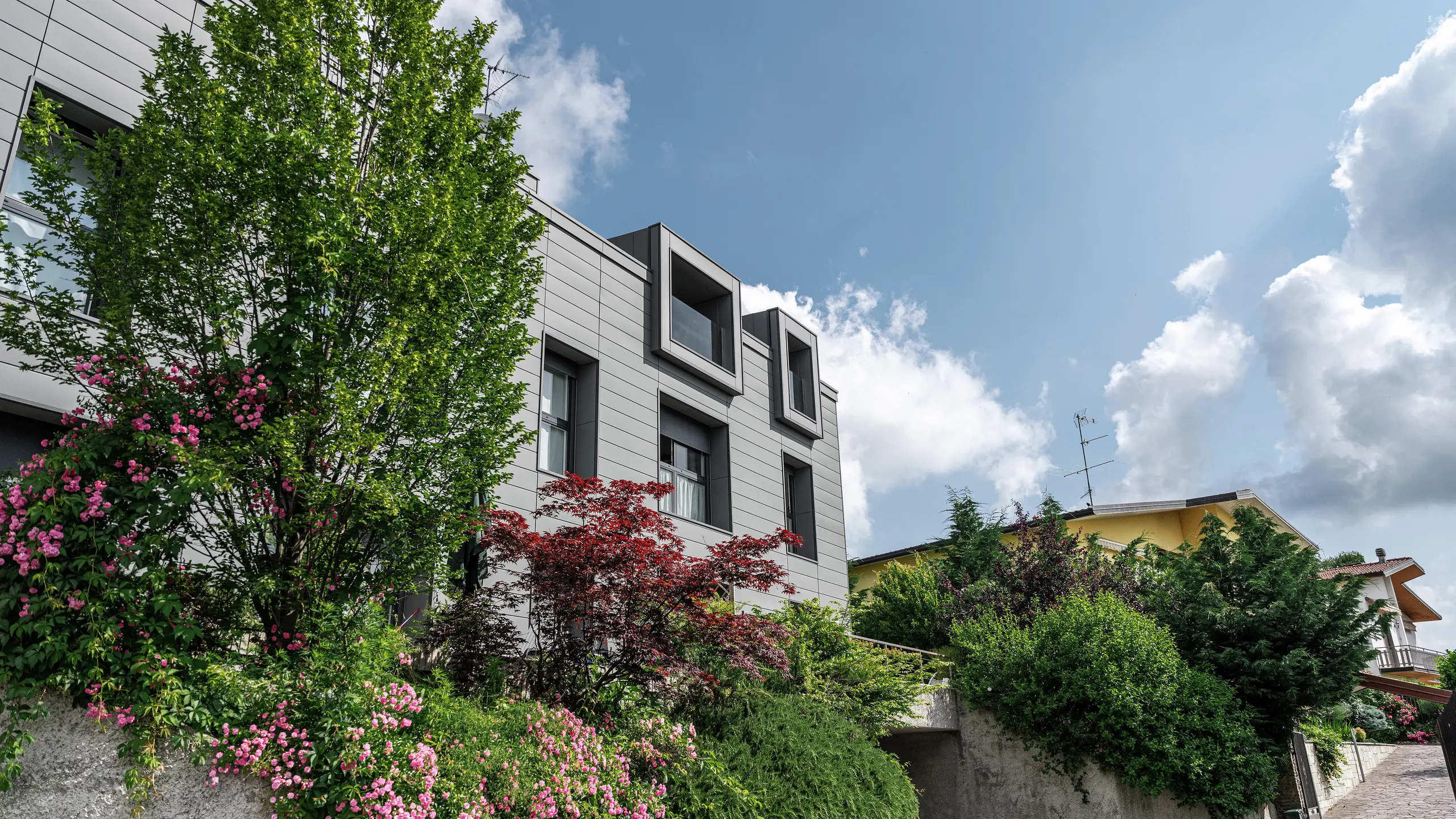 Moderní obytný komplex ve Faloppio v Itálii se stylovou fasádou z obkladů PREFA. Vyčnívající okna jsou jedinečným architektonickým prvkem, zatímco okolní rostliny a květiny podtrhují přirozenou krásu objektu. Odolné hliníkové obklady PREFA představují estetické a funkční řešení za každého počasí.