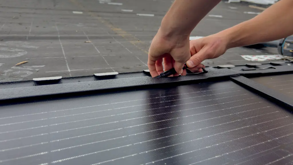 Detailní záběr na instalaci velkého integrovaného solárního panelu PREFA na střeše kostela sv. Karla Boromejského v bavorském Norimberku. Lesklé, obdélníkové panely jsou úhledně seřazeny vedle sebe a mají černý povrch. Na pravé straně obrazu jsou vidět ruce řemeslníka, jak zapojuje kabely solárního panelu. V pozadí je vidět střecha, která je již částečně nainstalována a připravena.
