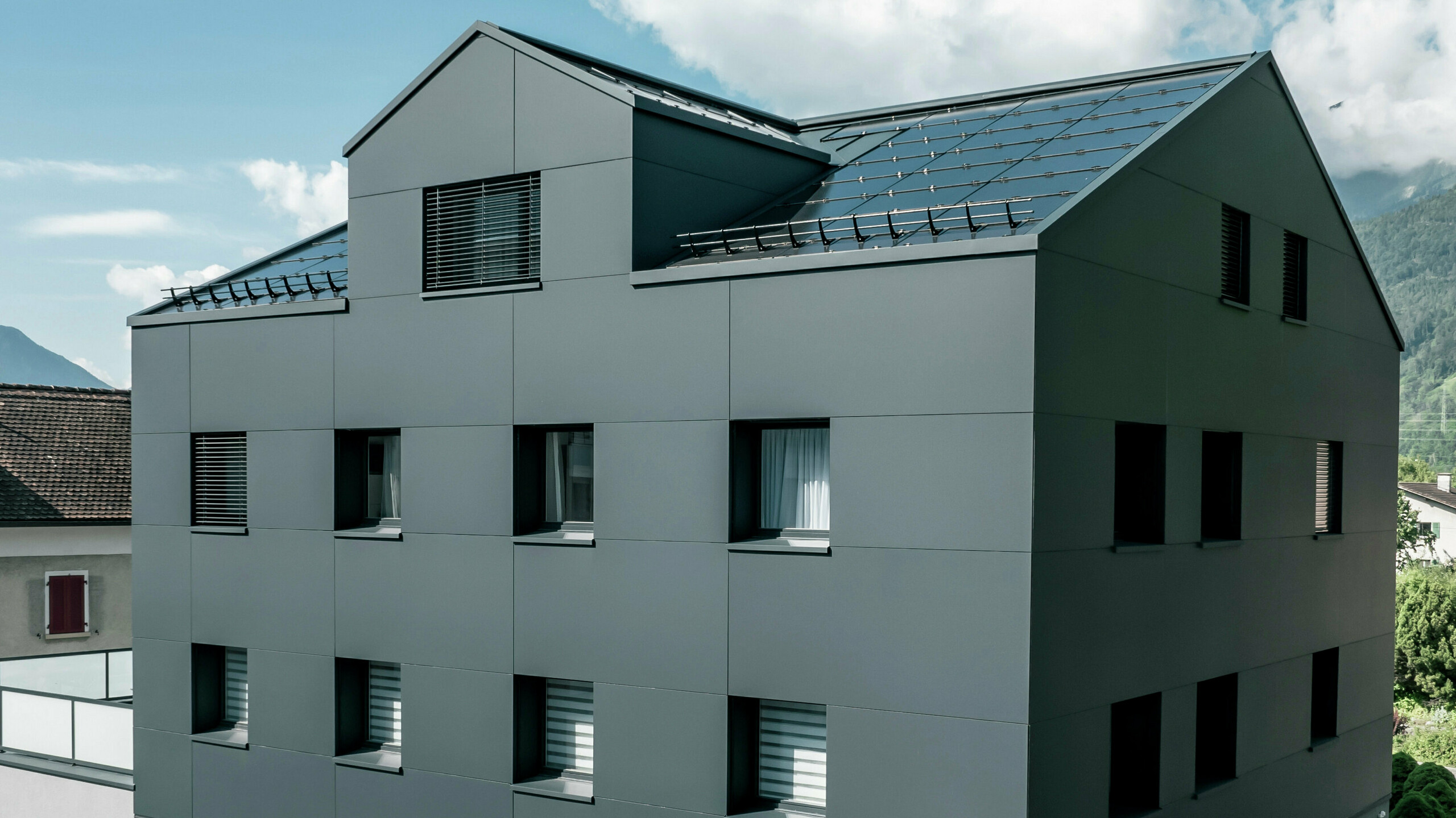 Modernisiertes Wohn- und Geschäftsgebäude nach der Fassadensanierung mit PREFABOND Aluminium Verbundplatten in Anthrazit. Die klare, elegante Linienführung der neuen Fassade verleiht dem Gebäude ein modernes Erscheinungsbild. Im Erdgeschoss befindet sich das Architekturbüro Voser. Die Umgebung mit Bäumen und Bergen rundet die harmonische Optik ab.