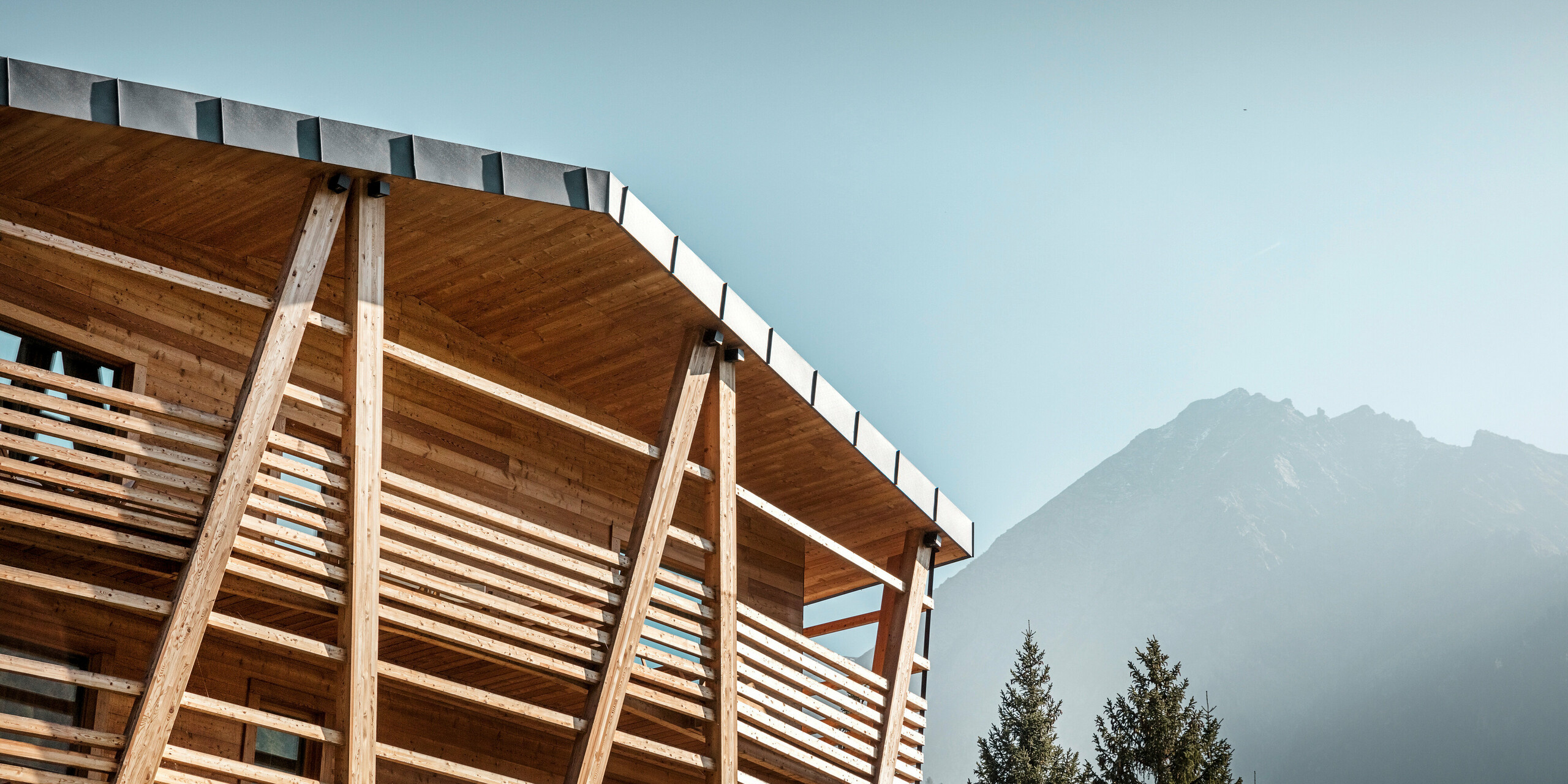 Nahaufnahme des Hotels 'Au Charmant Petit Lac', das mit seinen sichtbaren Holzstrukturen und dem darüber liegenden PREFALZ Aluminiumdach in P.10 Steingrau vor einer beeindruckenden Bergkulisse steht. Die Dachkante zeigt die präzise Verarbeitung der Stehfalzdeckung, die für ihre Langlebigkeit und Widerstandsfähigkeit gegen die alpinen Wetterbedingungen bekannt ist. Der Kontrast zwischen der natürlichen Holzfassade und dem metallischen Glanz des Daches betont das umweltfreundliche Design und die anspruchsvolle Architektur des Gebäudes.