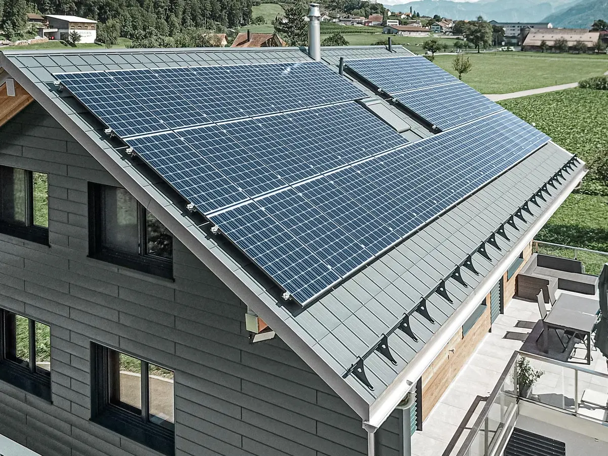 Obrázek ukazuje moderní rodinný dům s několika hliníkovými produkty PREFA. Velký fotovoltaický (FV) systém na střeše zdůrazňuje hodnotu udržitelné výroby energie. FV moduly jsou pečlivě instalovány po celé ploše střechy, aby se maximalizovalo využití solární energie. Integrovaný systém sněhových zábran, speciálně navržený pro použití se solárními panely, zabraňuje sesuvu sněhu ze střechy a chrání tak před lavinami. Na střeše byly instalovány osvědčené střešní panely PREFA v antracitové barvě. Tento robustní hliníkový střešní systém je obzvláště odolný a trvanlivý. Štítová část domu je obložena vysoce kvalitním obložením PREFA ve stejné barvě. Kombinace těchto prémiových kovových obkladů nabízí robustní ochranu i působivý vzhled.