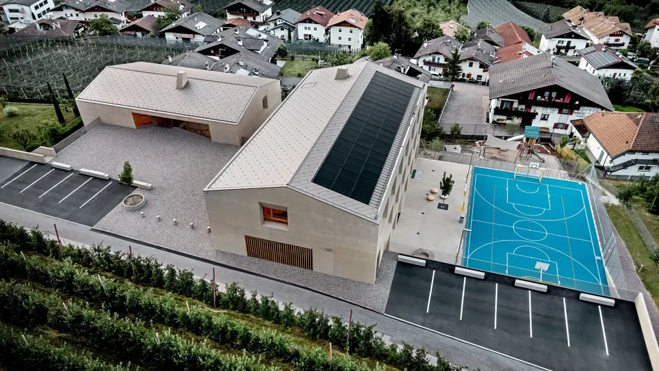 Letecký pohled na vzdělávací centrum Riffian v Jižním Tyrolsku s mateřskou školou, školou a sportovním hřištěm. Falcované šablony PREFA o rozměrech 44 x 44 cm v barvě P.10 bronzové tvoří precizní a odolnou hliníkovou střešní krytinu, která harmonicky zapadá do alpského prostředí. Moderní architektura se vyznačuje odolností proti povětrnostním vlivům a čistými designovými detaily.
