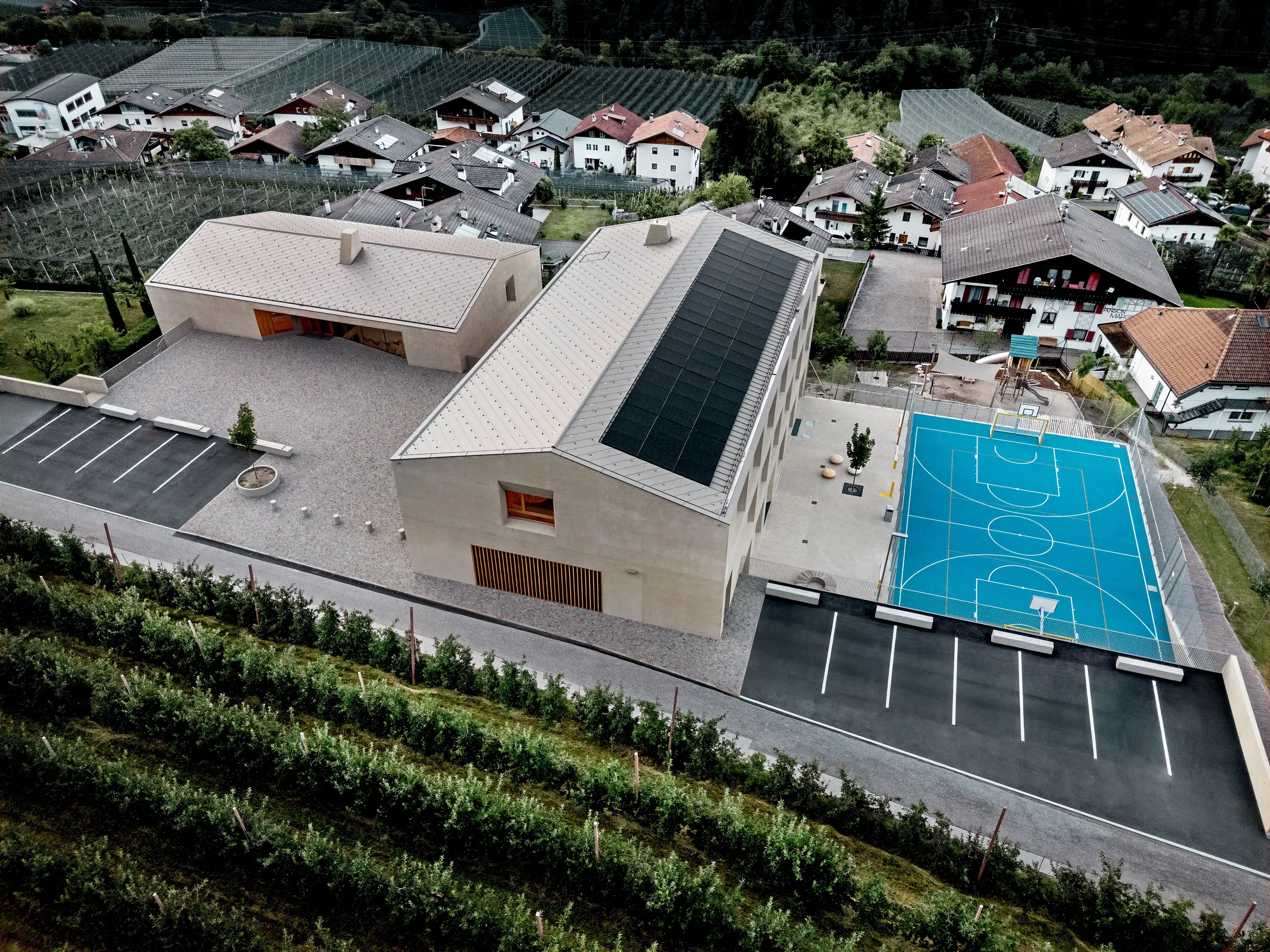 Letecký pohled na vzdělávací centrum Riffian v Jižním Tyrolsku s mateřskou školou, školou a sportovním hřištěm. Falcované šablony PREFA o rozměrech 44 x 44 cm v barvě P.10 bronzové tvoří precizní a odolnou hliníkovou střešní krytinu, která harmonicky zapadá do alpského prostředí. Moderní architektura se vyznačuje odolností proti povětrnostním vlivům a čistými designovými detaily.