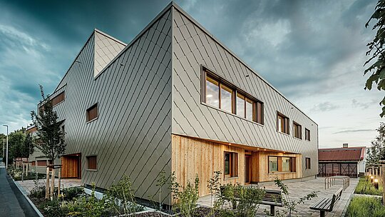 V jemném večerním světle se hybridní budova denní péče v Alzenau plně rozvíjí ve svém architektonickém působení. Výrazná fasáda, vyrobená z bronzových stěnových panelů PREFA 44 x 44, dodává budově elegantní, povětrnostním vlivům odolnou skořápku s jasnou strukturou. V přízemí vytváří vertikální dřevěné obložení přirozený, teplý kontrast k hliníku a zdůrazňuje vstupní prostor i chráněná místa k sezení. Minimalistický, moderní design ladí s udržovaným areálem, kde lavičky, stojany na kola a mladé stromy zvou návštěvníky k posezení. Obrázek působivě ukazuje, jak odolné produkty PREFA přispívají k udržitelné, funkční a příjemné vzdělávací budově.