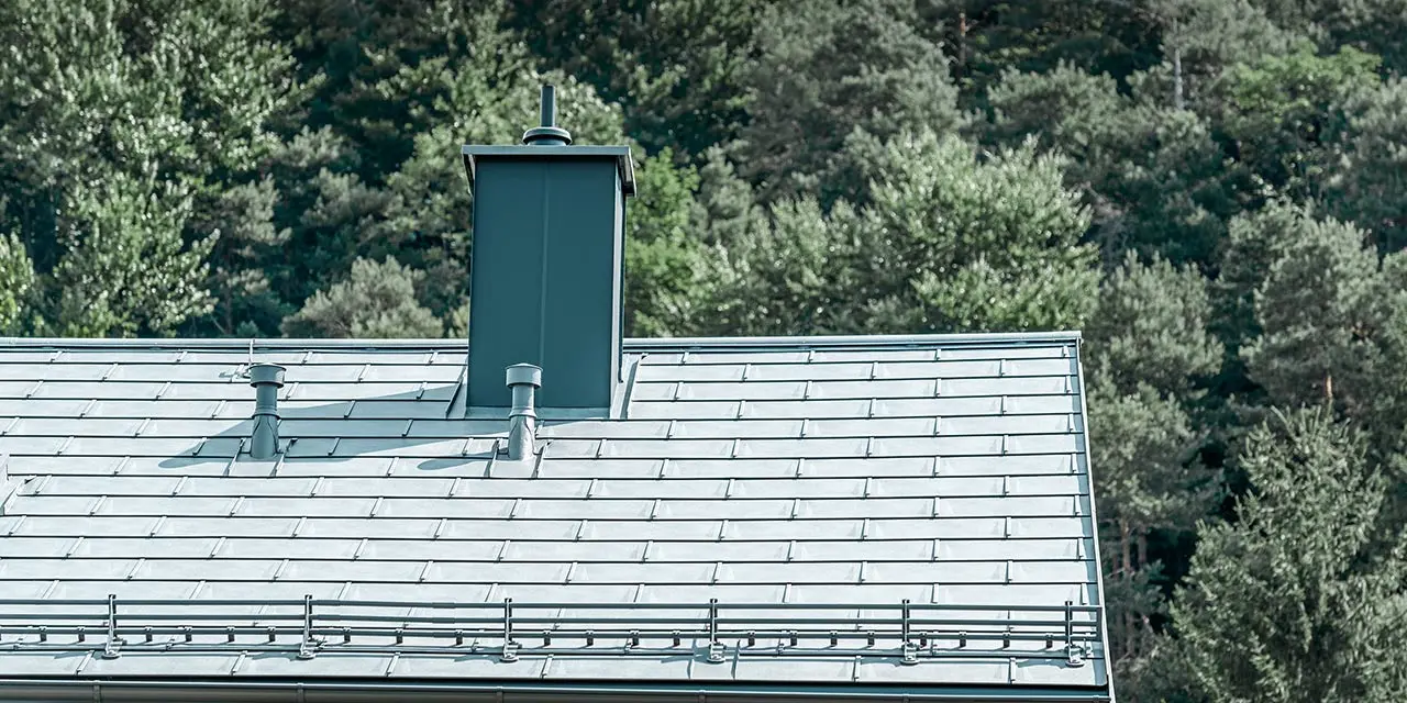 Detailní pohled na sedlovou střechu pokrytou střešním panelem PREFA R.16 v barvě P.10 břidlicové se systémem pro zadržování sněhu, oplechováním komína, okapem a odvětrávací trubkou, vše od PREFA