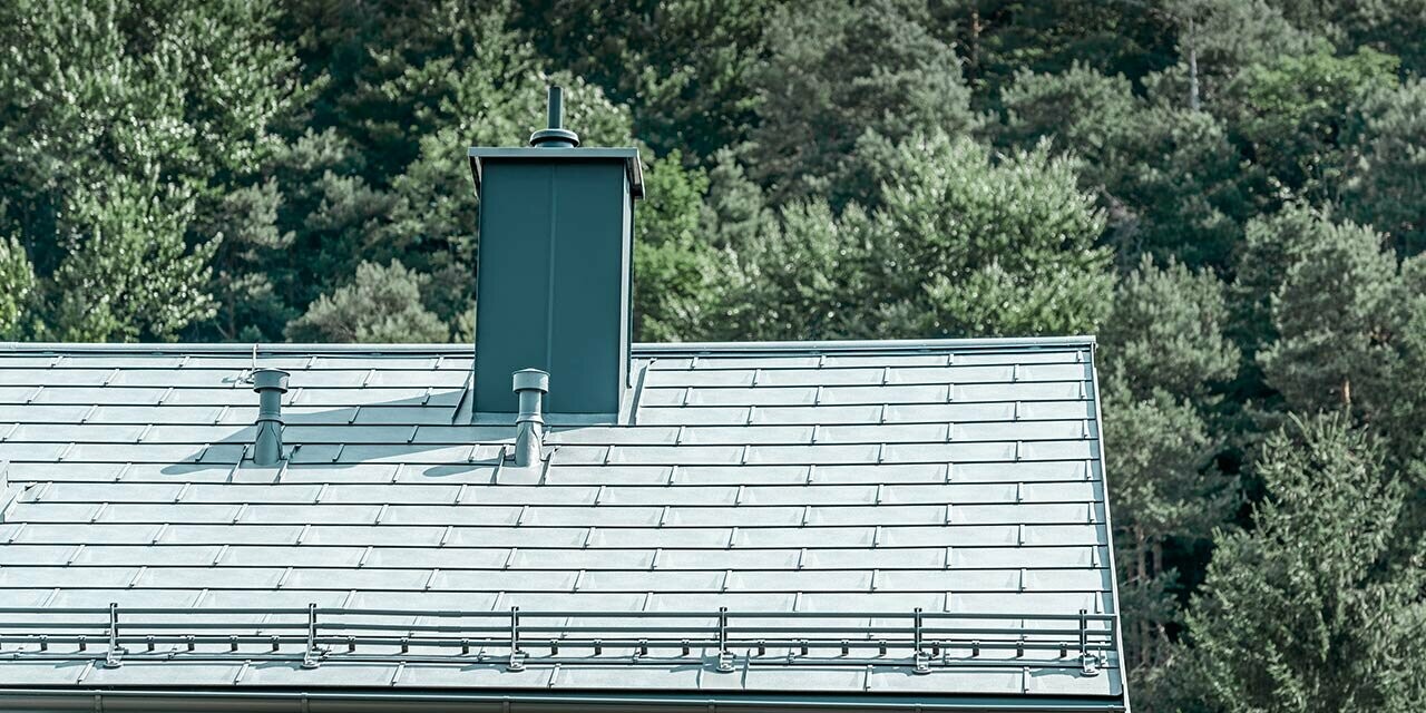 Detailní pohled na sedlovou střechu pokrytou střešním panelem PREFA R.16 v barvě P.10 břidlicové se systémem pro zadržování sněhu, oplechováním komína, okapem a odvětrávací trubkou, vše od PREFA
