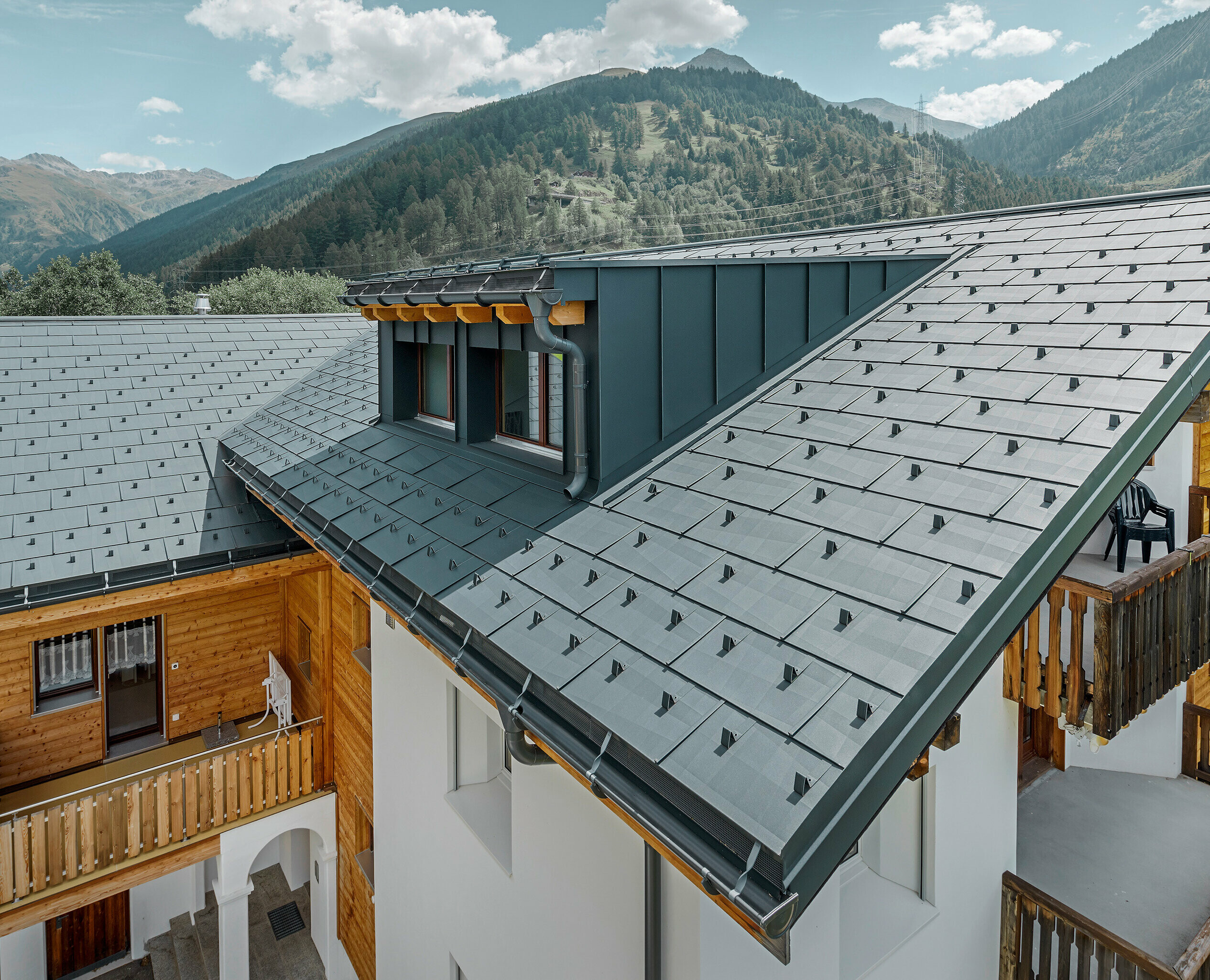 Bytový dům s vikýřem a sedlovou střechou pokrytou střešními panely PREFA FX.12 v antracitové barvě; v pozadí se vypínají hory;