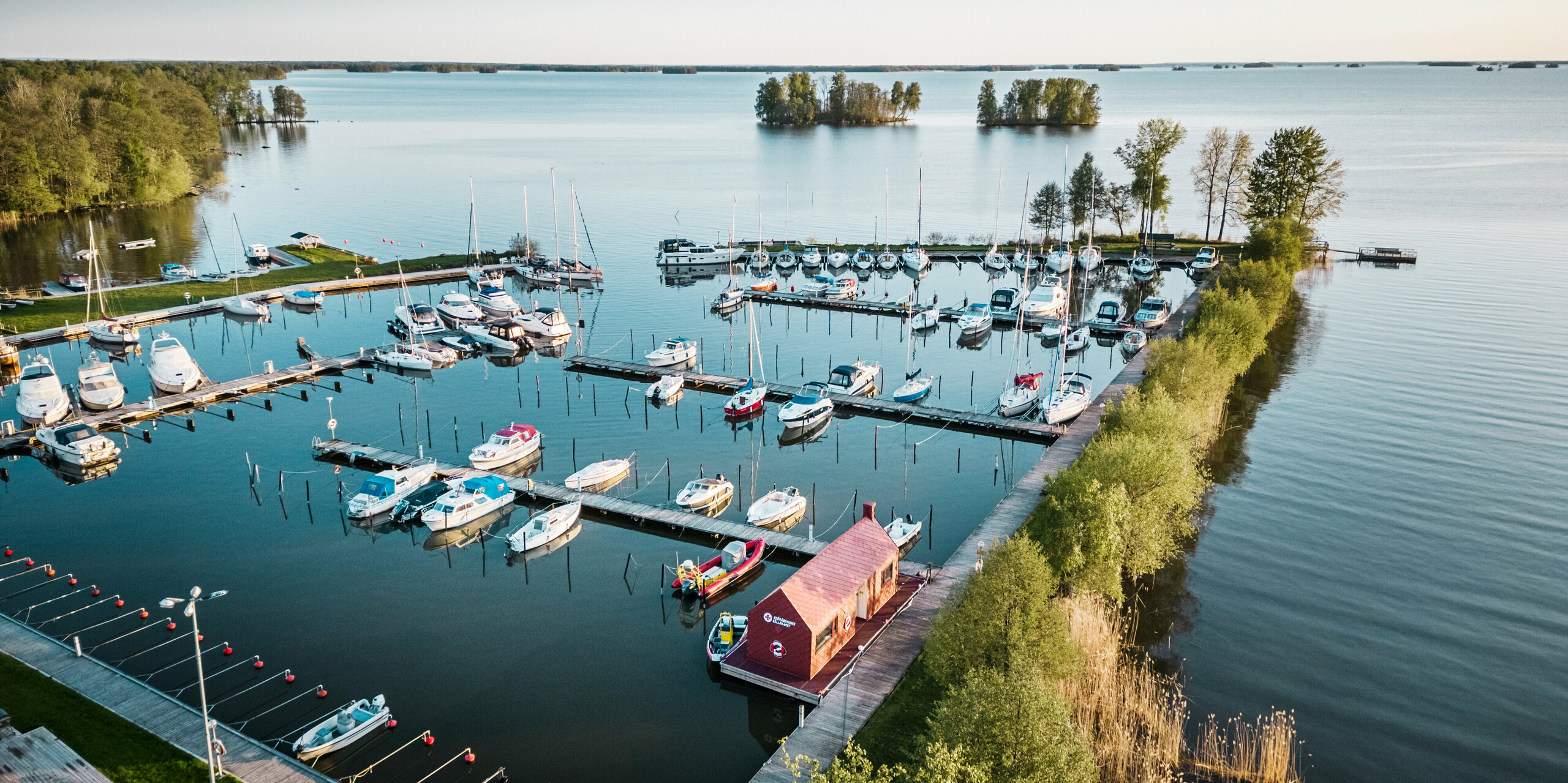 Letecký pohled na námořní záchranný dům v přístavu na jezeře Hjälmaren ve Švédsku, uhnízděný mezi plachetnicemi a klidnou modrou vodou. Poutavá tmavě červená střecha a fasáda domu z falcovaných a fasádních šablon PREFA 29×29 harmonicky zapadají do mořského prostředí a zároveň vytvářejí silný vizuální akcent. Záchranná stanice slouží jako životně důležitá základna pro vodní záchranáře a je svědectvím o propojení člověka s přírodou a nutnosti chránit obojí.