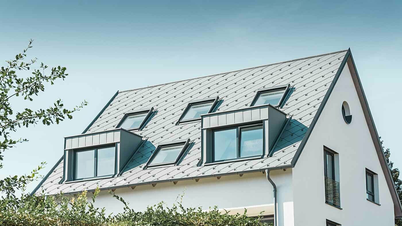 Klasická sedlová střecha se dvěma vikýři a několika střešními okny, pokrytá PREFA střešními šablonami 44 × 44 světle šedé barvy.